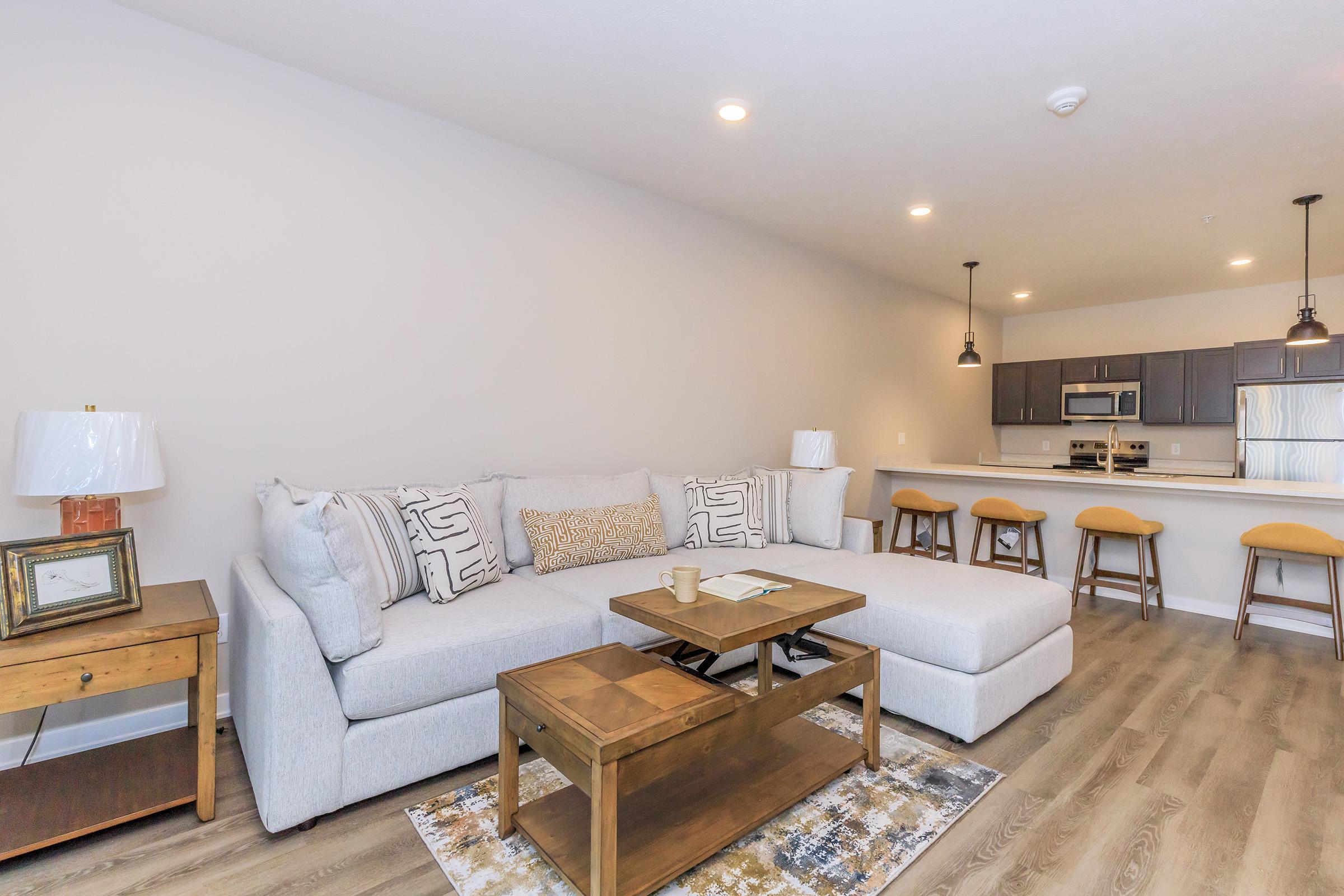 A modern living room featuring a light gray sectional sofa with decorative cushions, a wooden coffee table, and a lamp on a side table. In the background, there’s a kitchen area with dark cabinetry and bar stools, creating an open-concept design with soft lighting and wooden flooring.