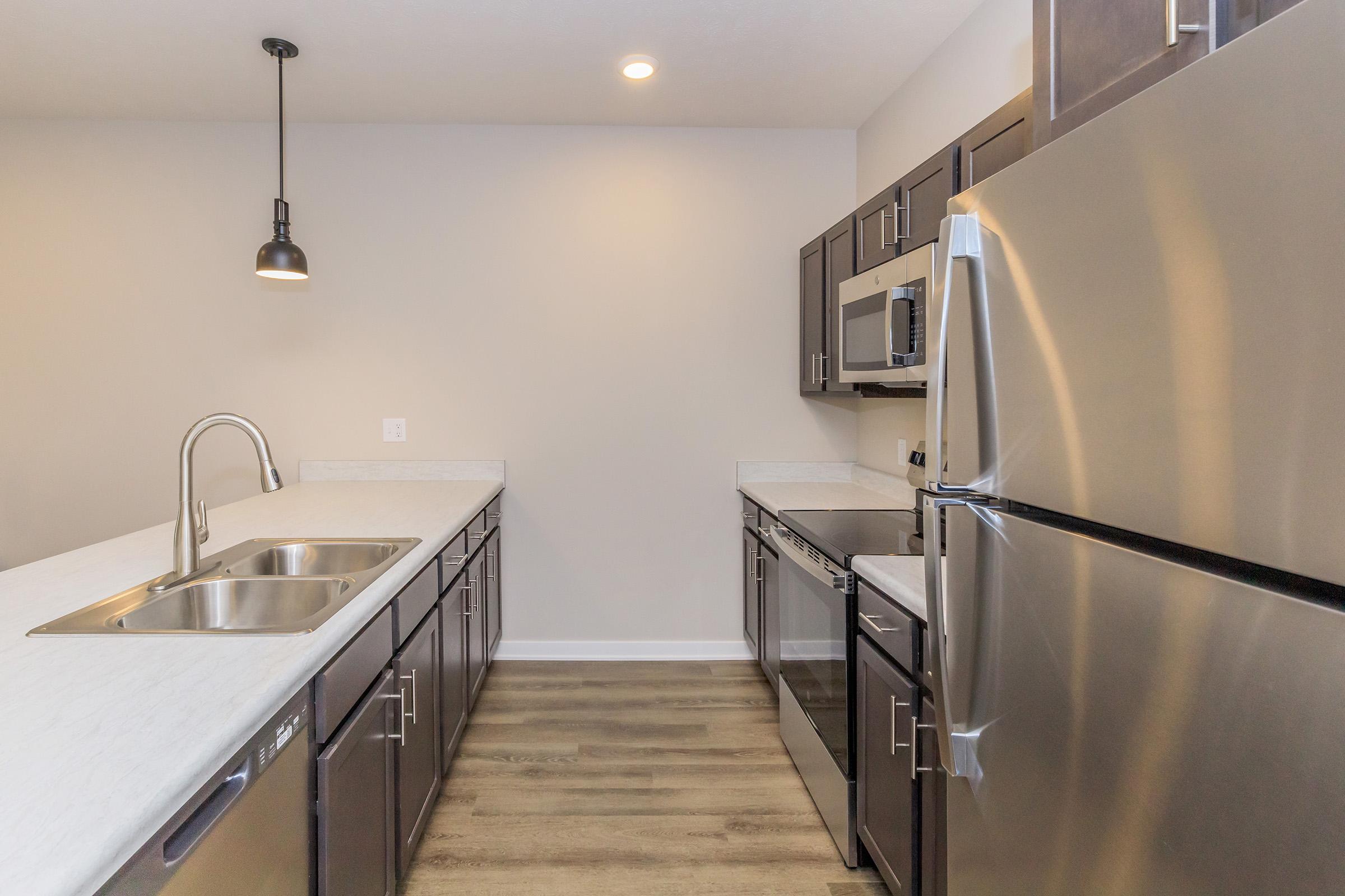 Modern kitchen featuring dark wood cabinets, stainless steel appliances, a double sink, and a neutral-colored wall. The space includes a microwave, dishwasher, and a sleek countertop. A pendant light hangs above the kitchen island, and the flooring is a light-colored wood laminate.