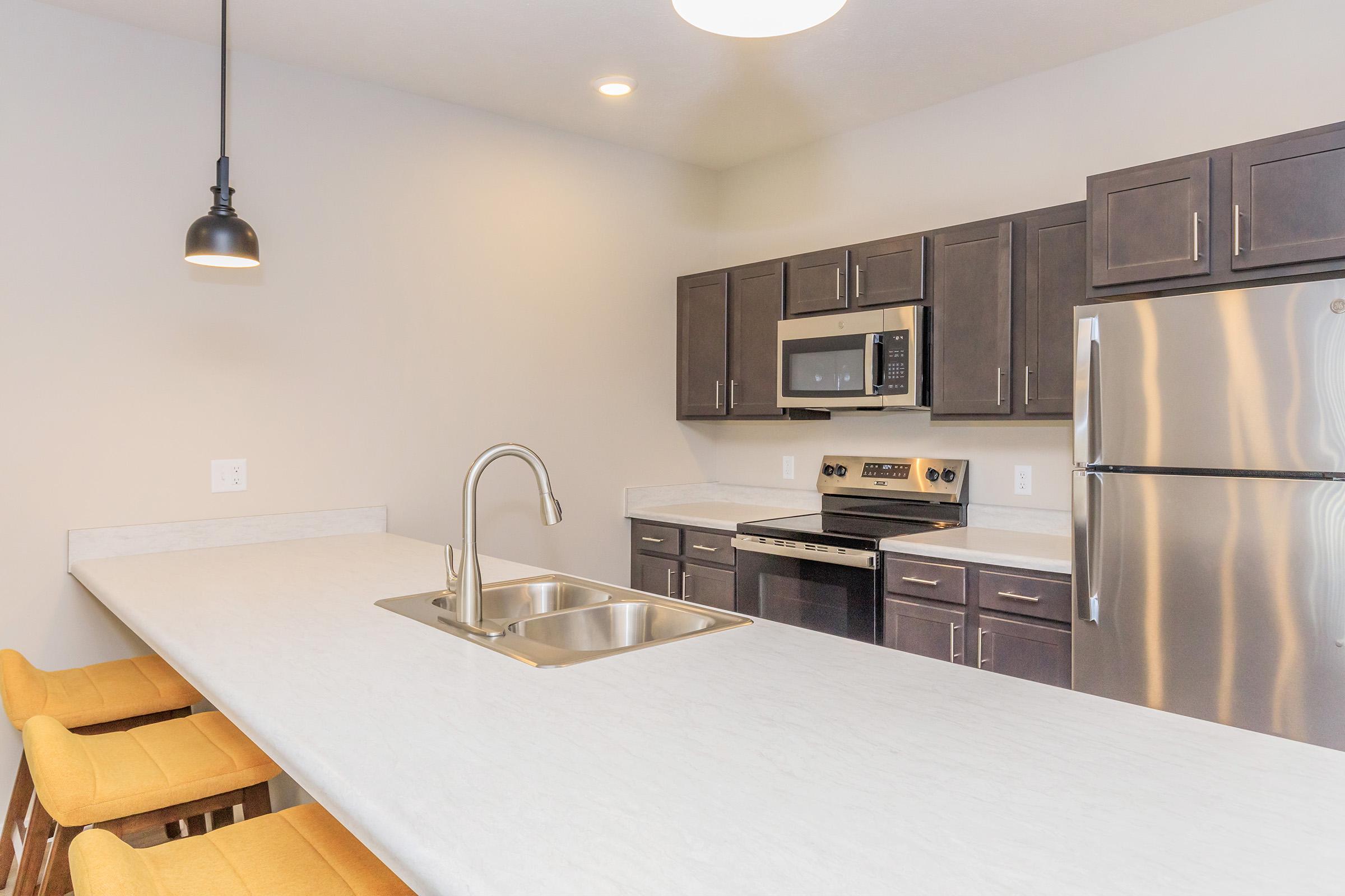 Modern kitchen featuring dark wooden cabinets, stainless steel appliances, and a spacious island with a double sink. Brightly lit with a pendant light, the kitchen includes a stainless steel fridge and an oven. Yellow bar stools are positioned at the island, creating a welcoming atmosphere.