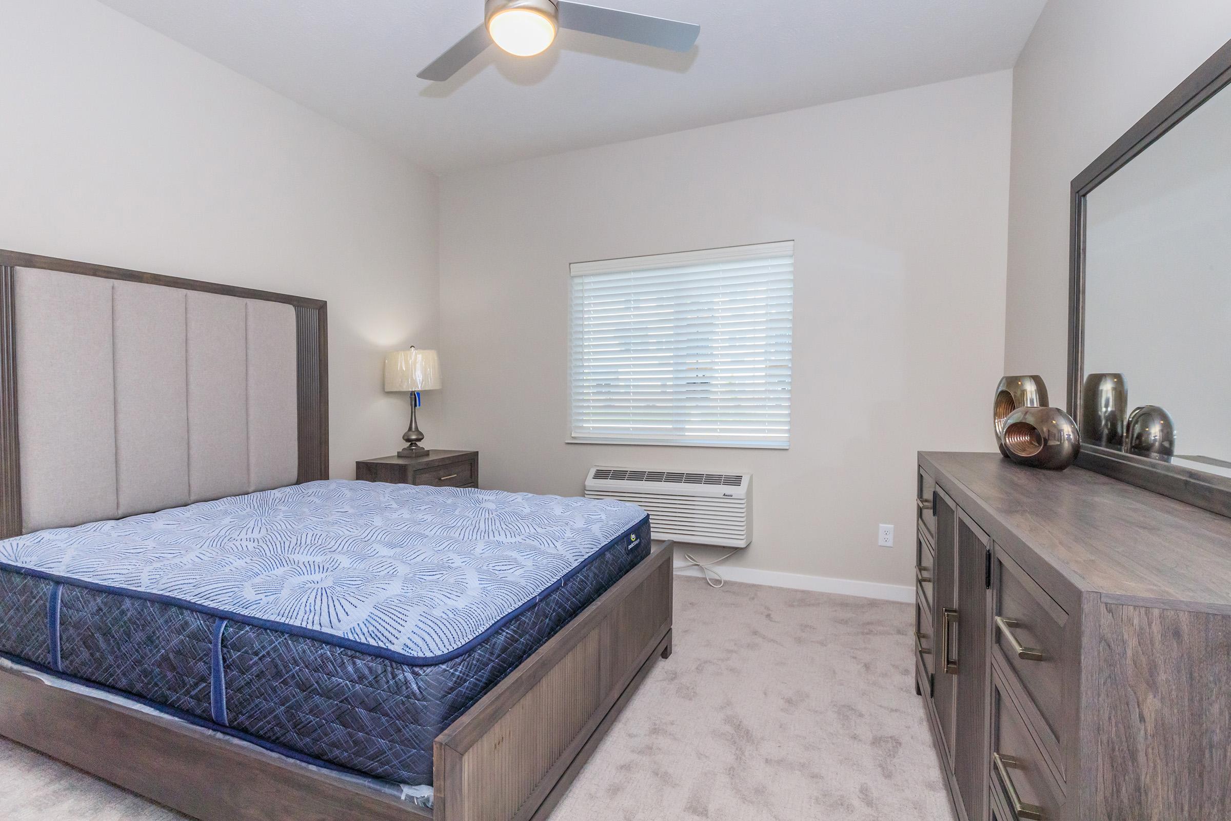 A modern bedroom featuring a large bed with a blue mattress, a stylish headboard, a nightstand with a lamp, and a dresser. There is an air conditioning unit beneath a window with blinds, and the carpeted floor adds a cozy touch to the neutral-colored walls.