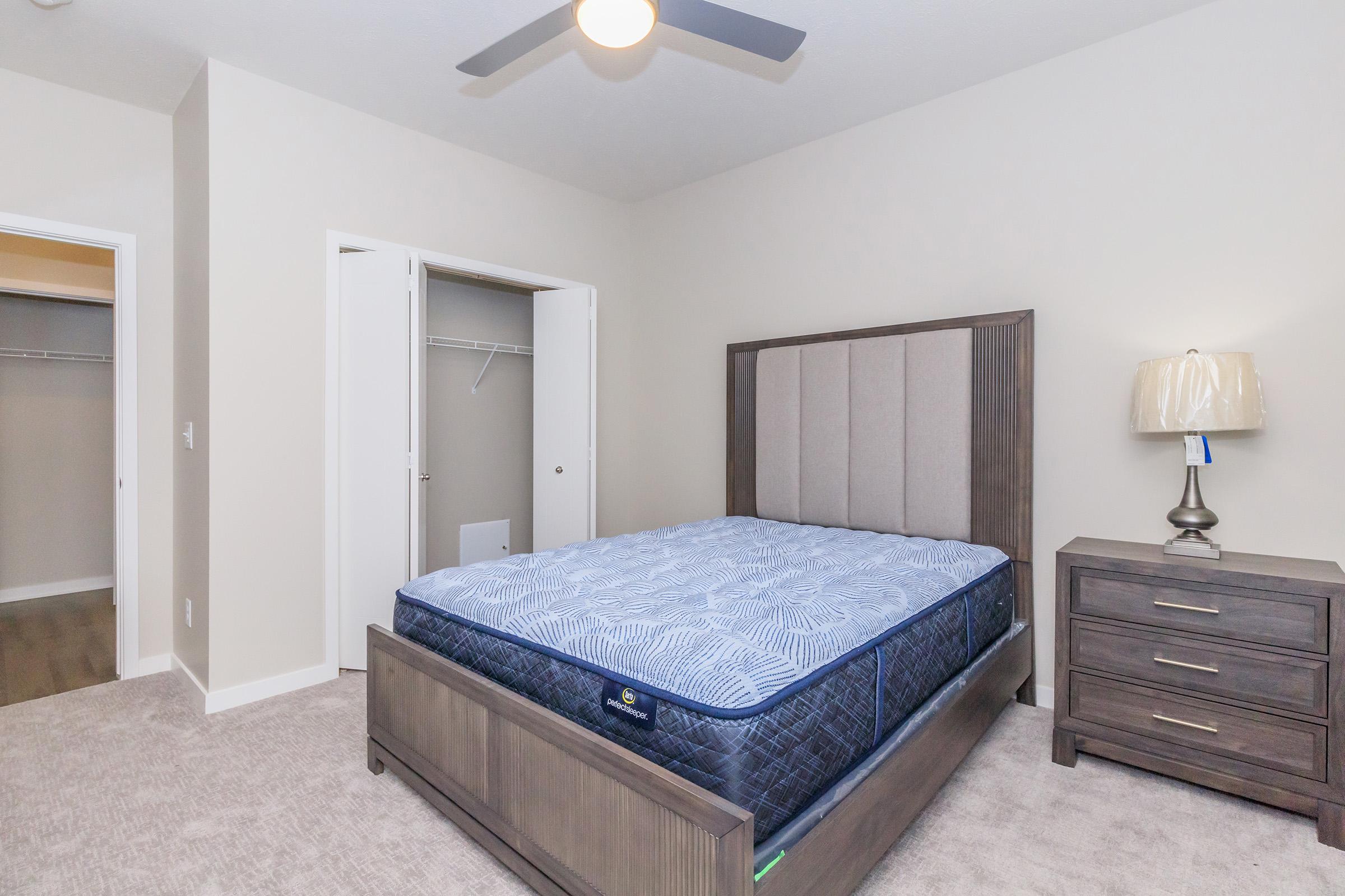A modern bedroom featuring a large bed with a gray upholstered headboard, a bedside lamp on a dark wooden nightstand, and a beige wall color. There’s a closet with sliding doors and a light-colored carpeted floor, creating a clean and inviting space.
