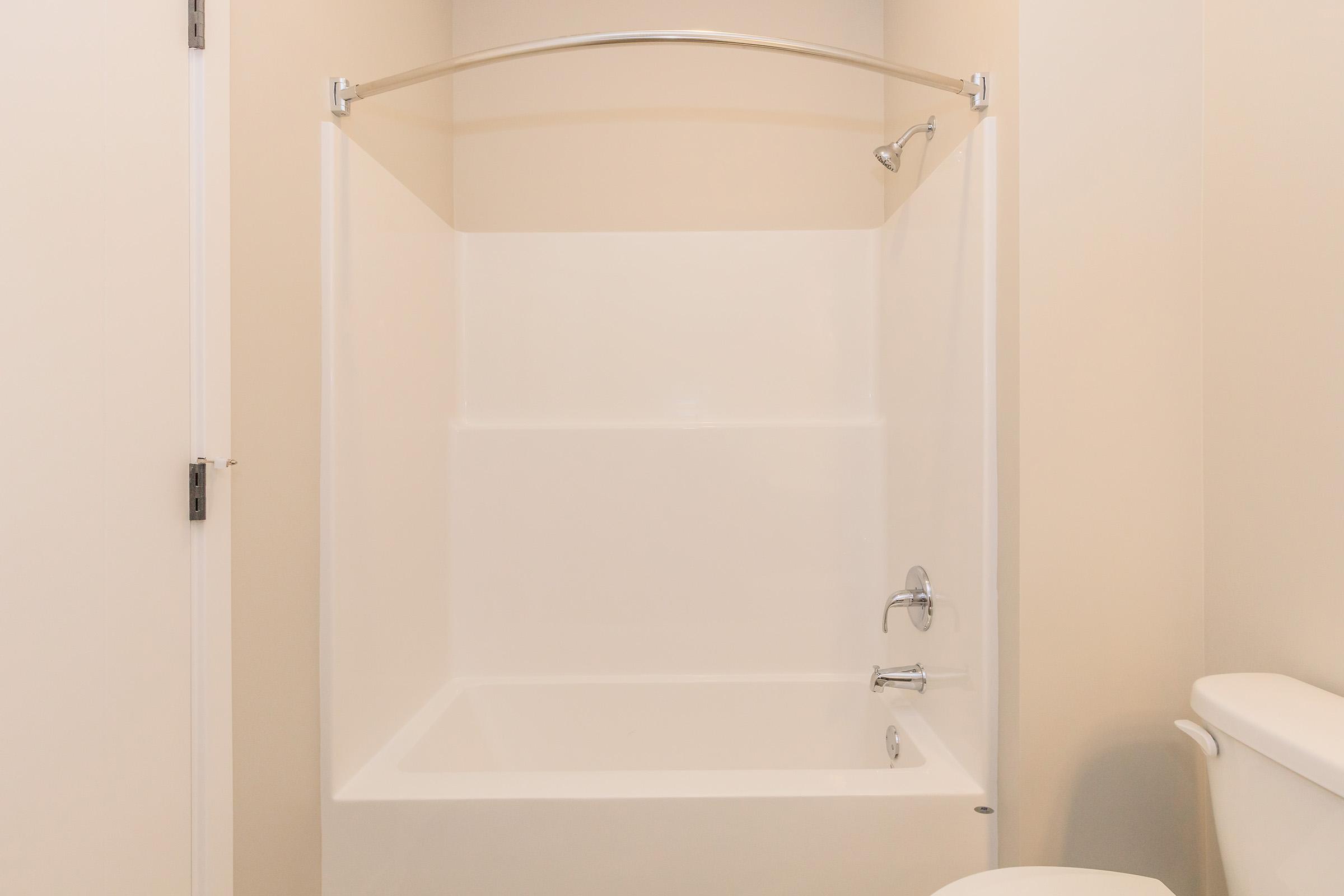 Empty bathroom with a white shower tub and sliding glass door, light beige walls, and a white toilet nearby. The bathtub has a chrome faucet and showerhead. The overall space appears clean and minimalistic.