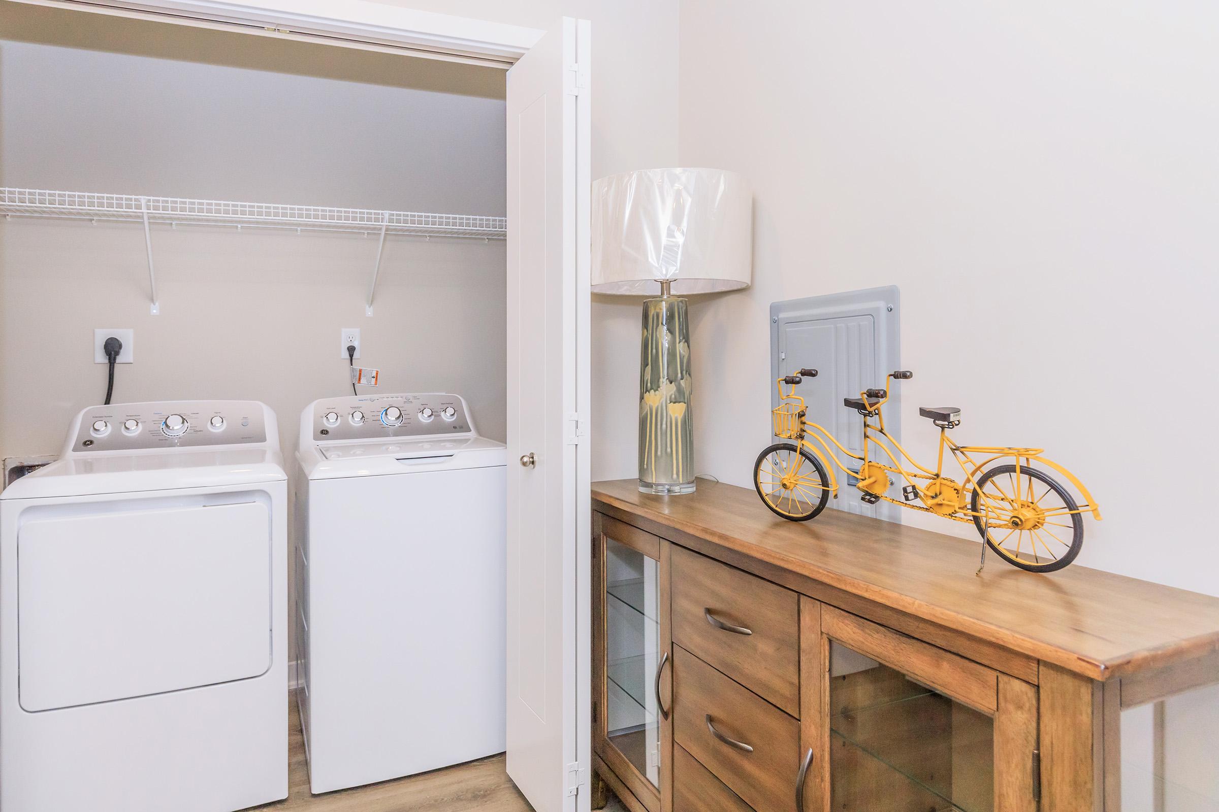 A laundry room featuring two front-loading washing machines, a wooden dresser with glass doors, a decorative yellow bicycle model, and a lamp on top of the dresser. The walls are painted a soft color, and there is a shelf above the machines for additional storage.