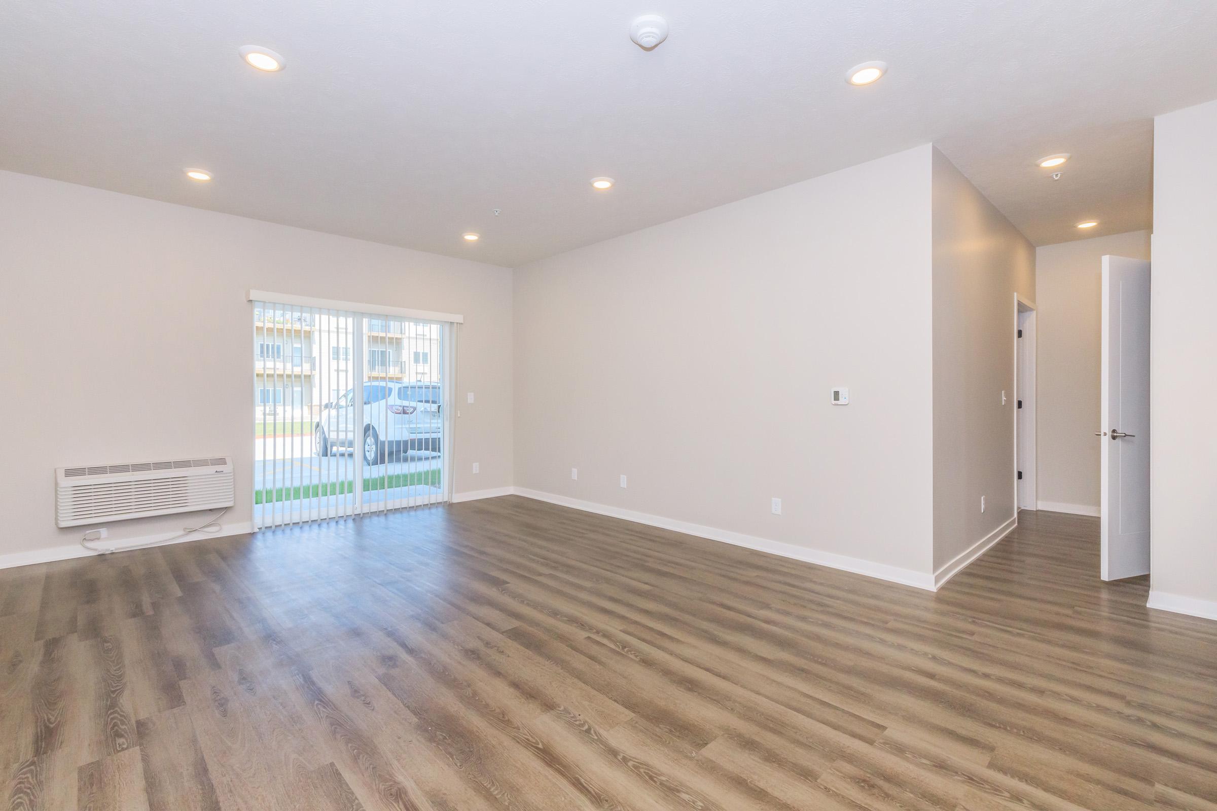 A spacious, unfurnished living room with light-colored walls and warm wooden flooring. Large windows allow natural light to enter, and a door leads to an outdoor area. Visible amenities include a wall-mounted air conditioning unit and internal doorways. The overall atmosphere is bright and open.