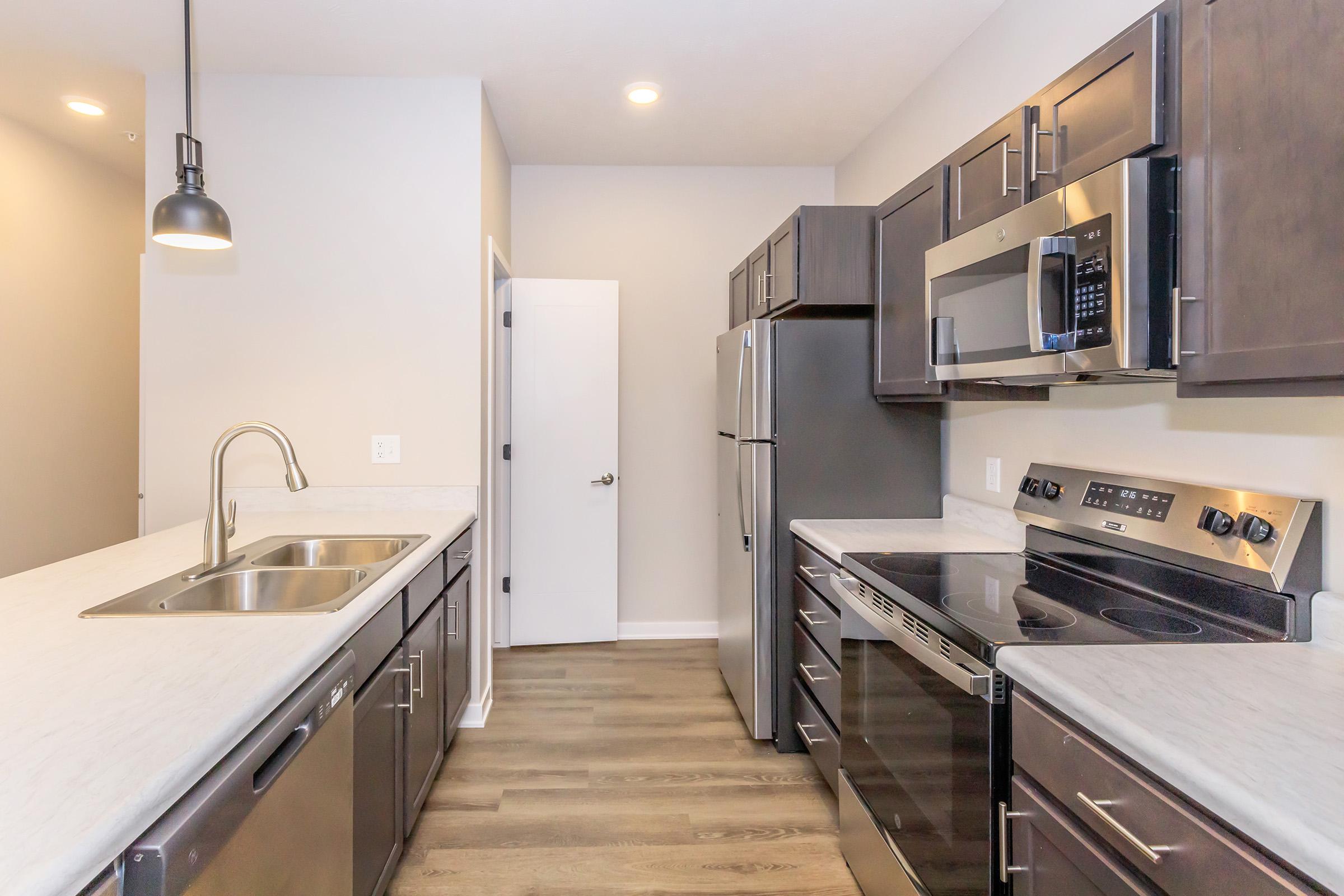 Modern kitchen featuring dark wood cabinets, stainless steel appliances including a refrigerator, microwave, and oven. There is a double sink with a sleek faucet and a light fixture hanging above. The countertop is a light-colored stone, and the floor is done in a wood-style finish. Bright and spacious atmosphere.