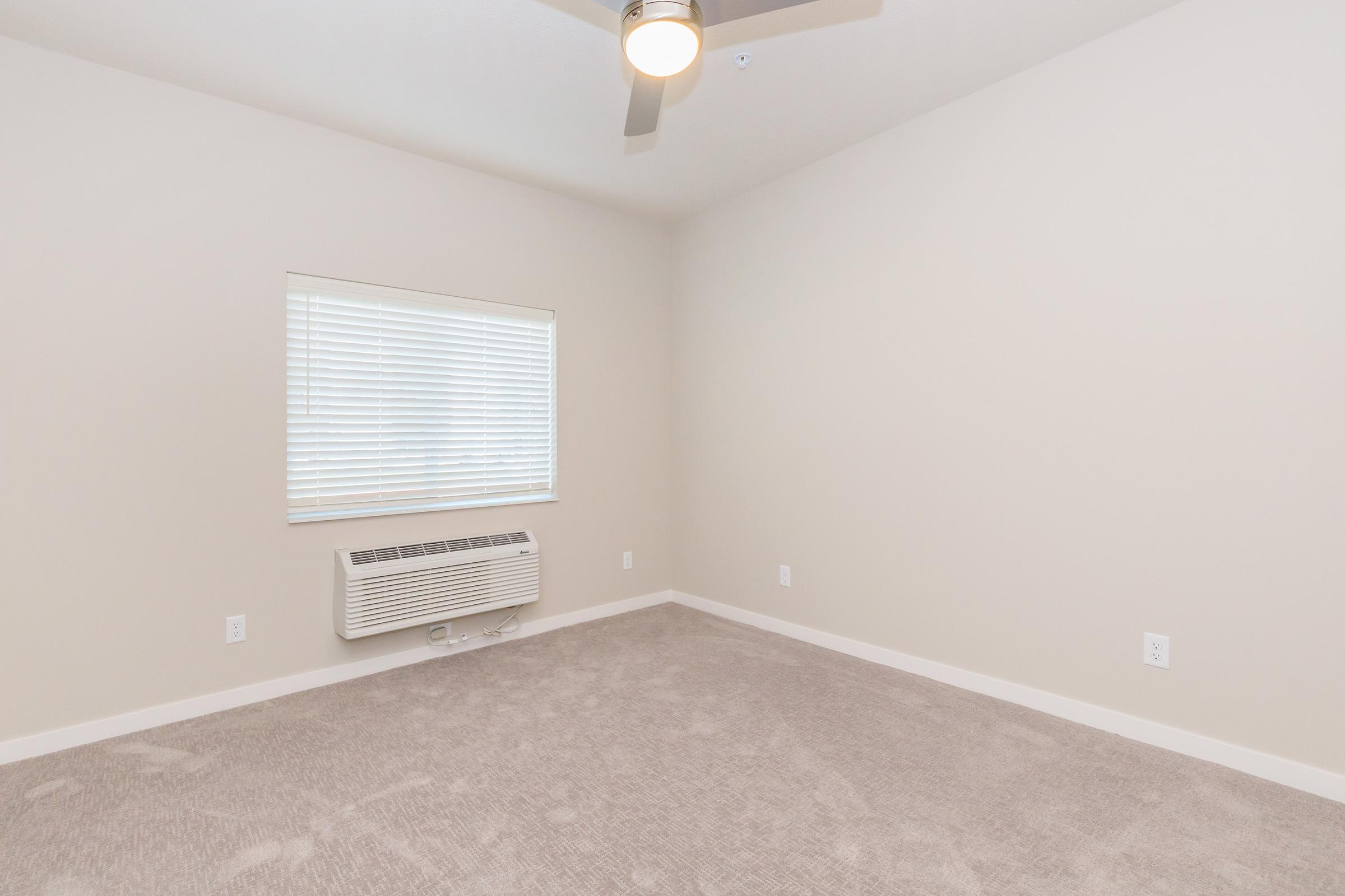 A spacious and empty bedroom with light beige walls and plush carpet. A window with white blinds allows natural light to enter, and a ceiling fan is mounted above. An air conditioning unit is visible on the wall below the window. The room is uncluttered and ready for furniture.