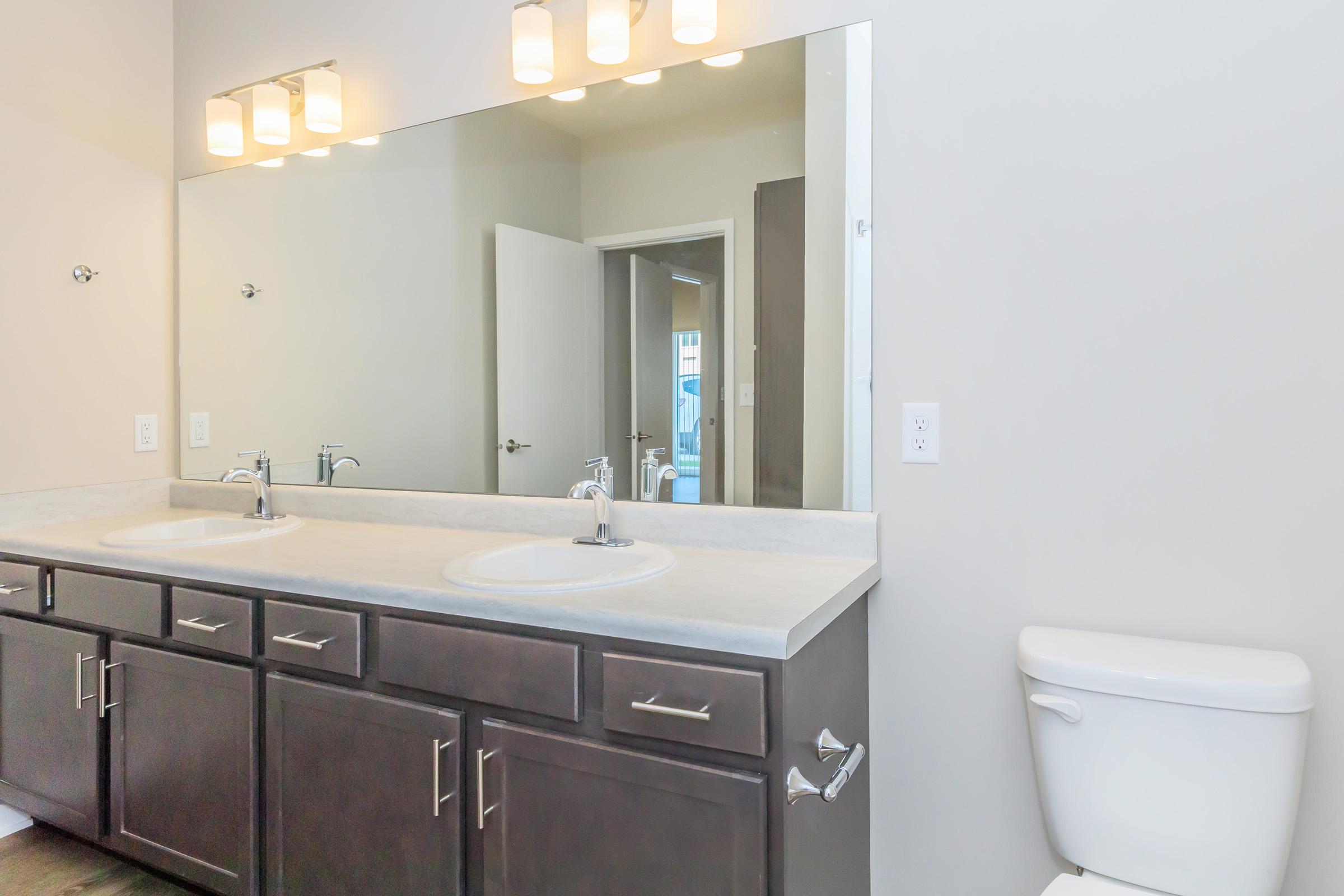 A modern bathroom featuring a double sink vanity with two circular sinks and a large mirror. The cabinets are dark wood, and there is a white toilet to the right. The walls are painted light gray, and the space is well-lit with overhead light fixtures. A doorway is visible in the background.