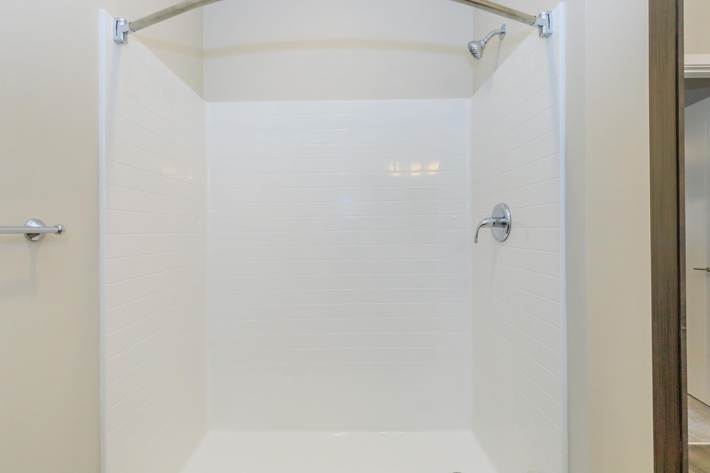 A clean, empty shower stall with white tiled walls and a chrome showerhead. The space is well-lit and has a minimalist design, featuring a curved shower rod at the top. The floor is also white and free of any objects or decor.