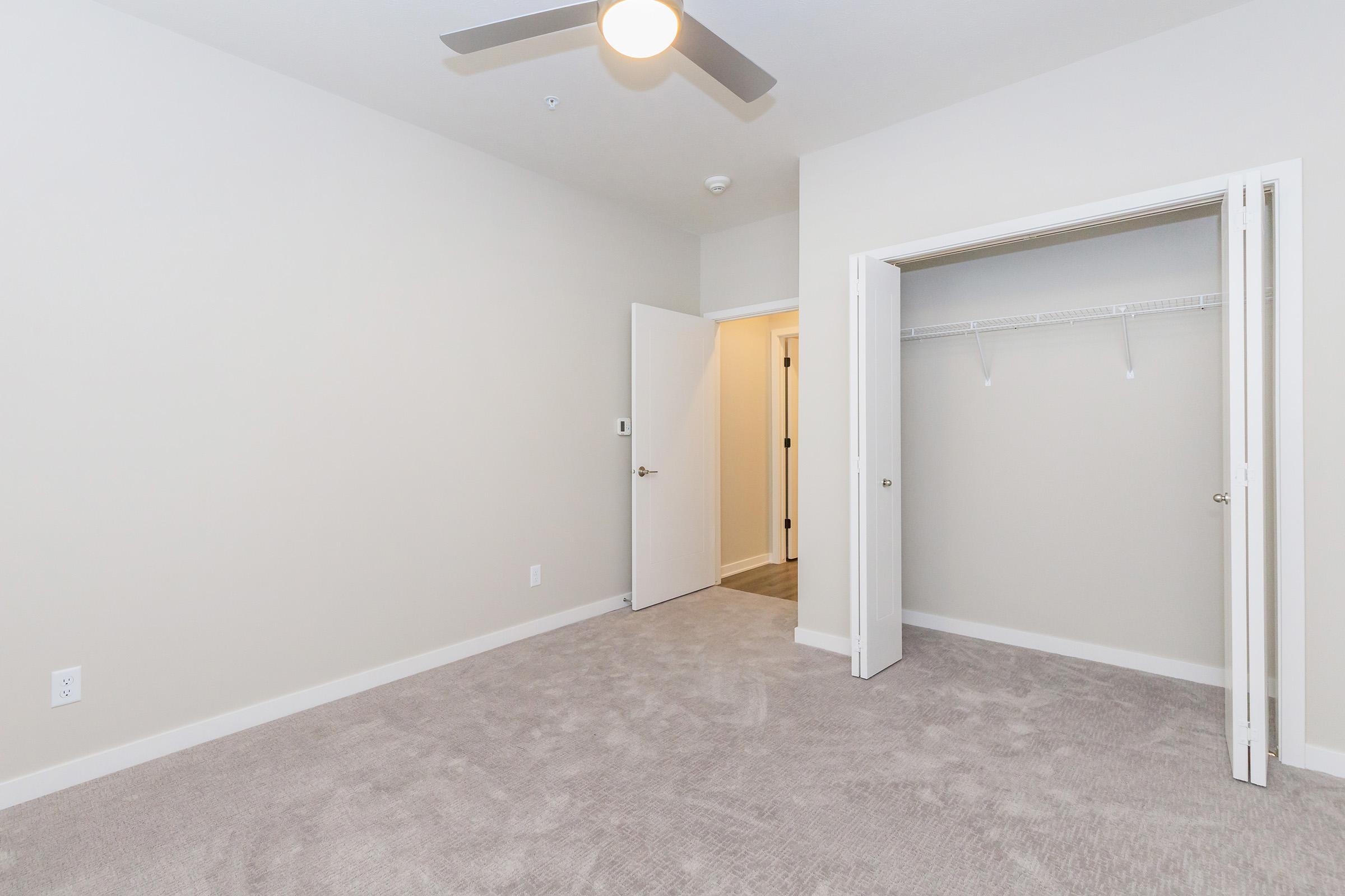 An empty room with light gray carpet, painted pale walls, and a ceiling fan. A doorway leads to another room, while a closet with double doors is visible on one wall. The space is well-lit and uncluttered, creating a bright and open feel.