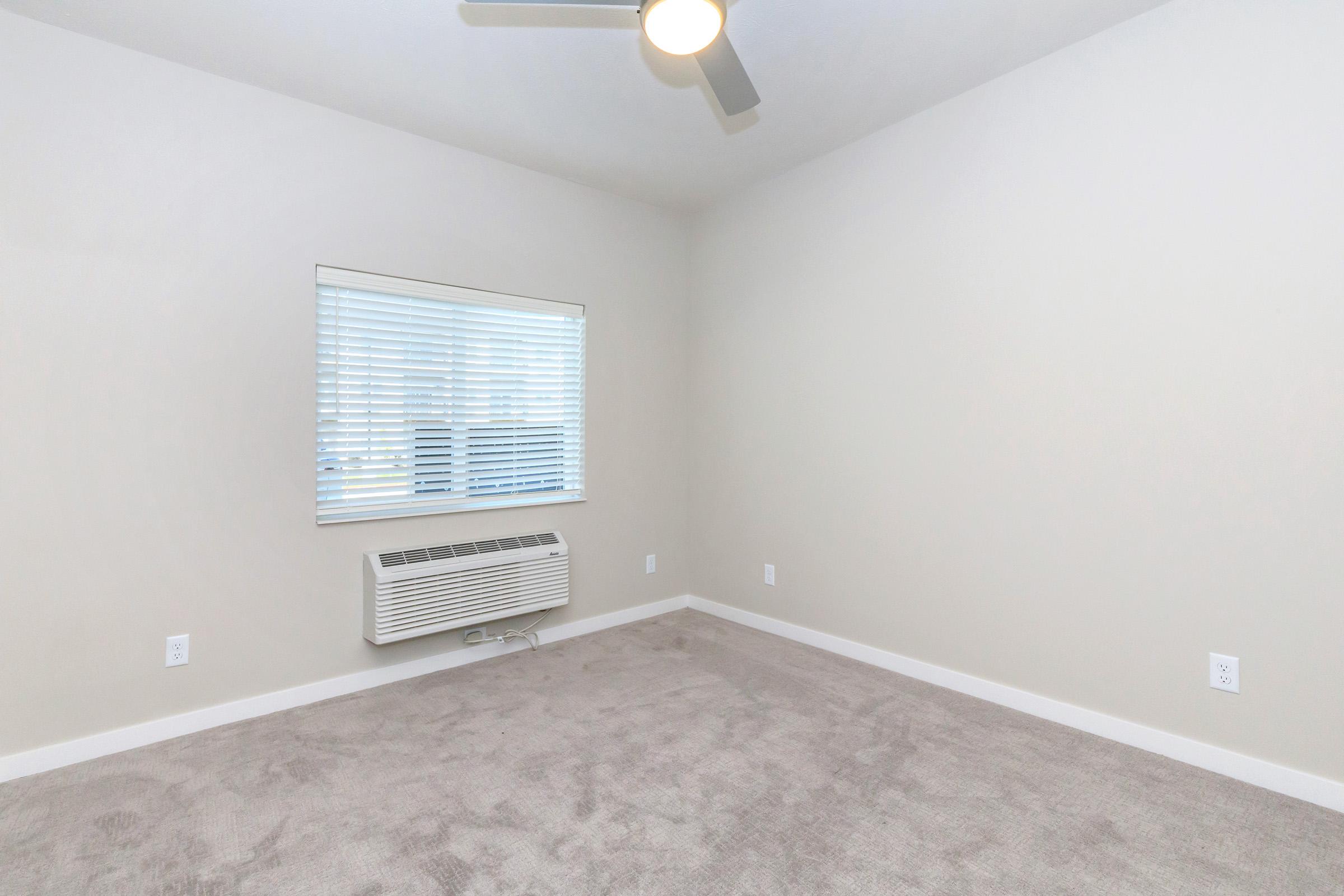 A vacant, minimally furnished room with light gray carpet and beige walls. The room features a ceiling fan, a window with blinds, and a wall-mounted air conditioning unit. The overall atmosphere is bright and neutral, suitable for various decorating styles.