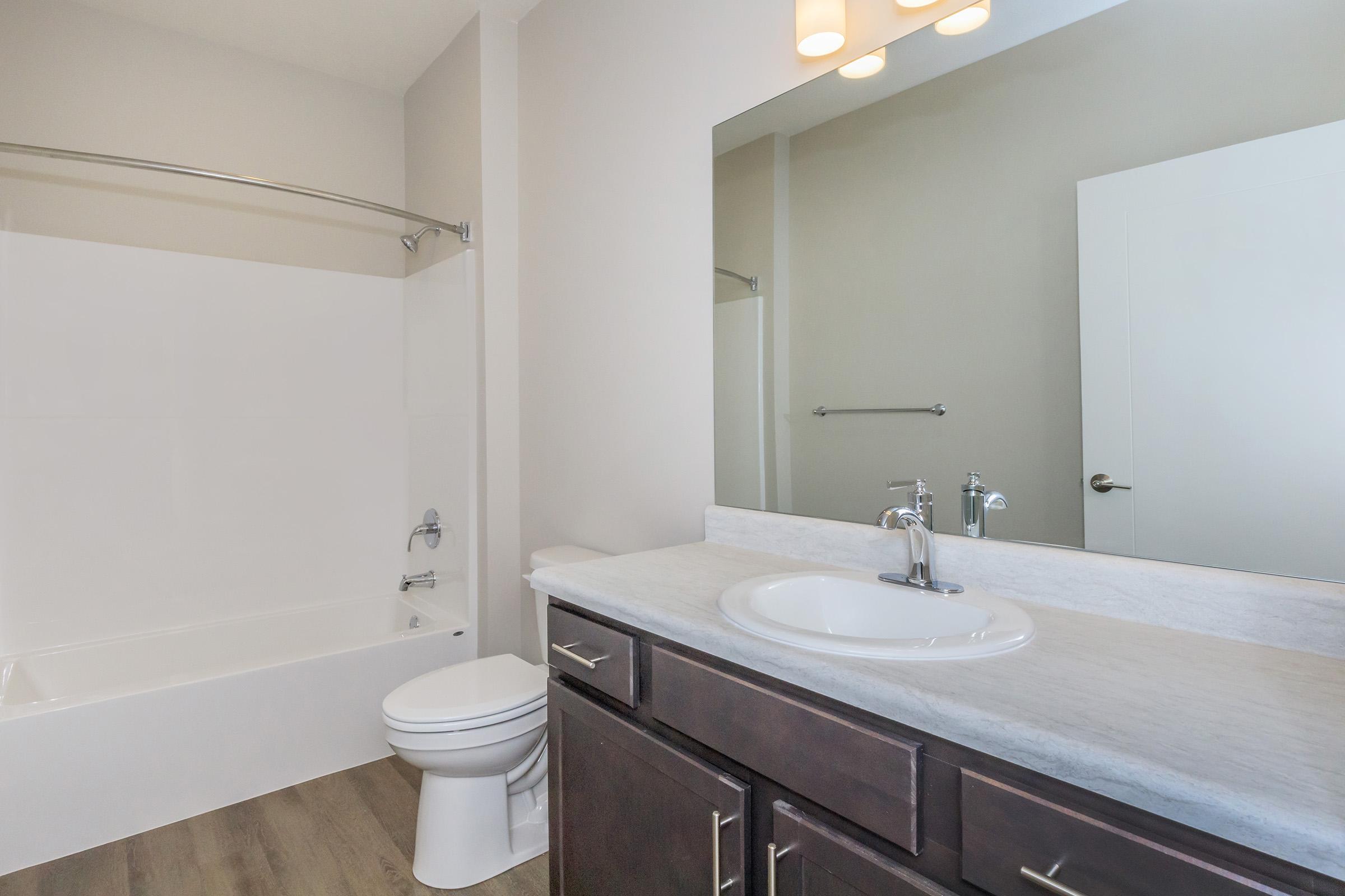 A clean and modern bathroom featuring a white bathtub and shower combination, a sink with a marble countertop, and a large mirror above. The walls are painted in neutral tones, and there is a single light fixture above the mirror. The floor has a wood-like finish, enhancing the contemporary design.