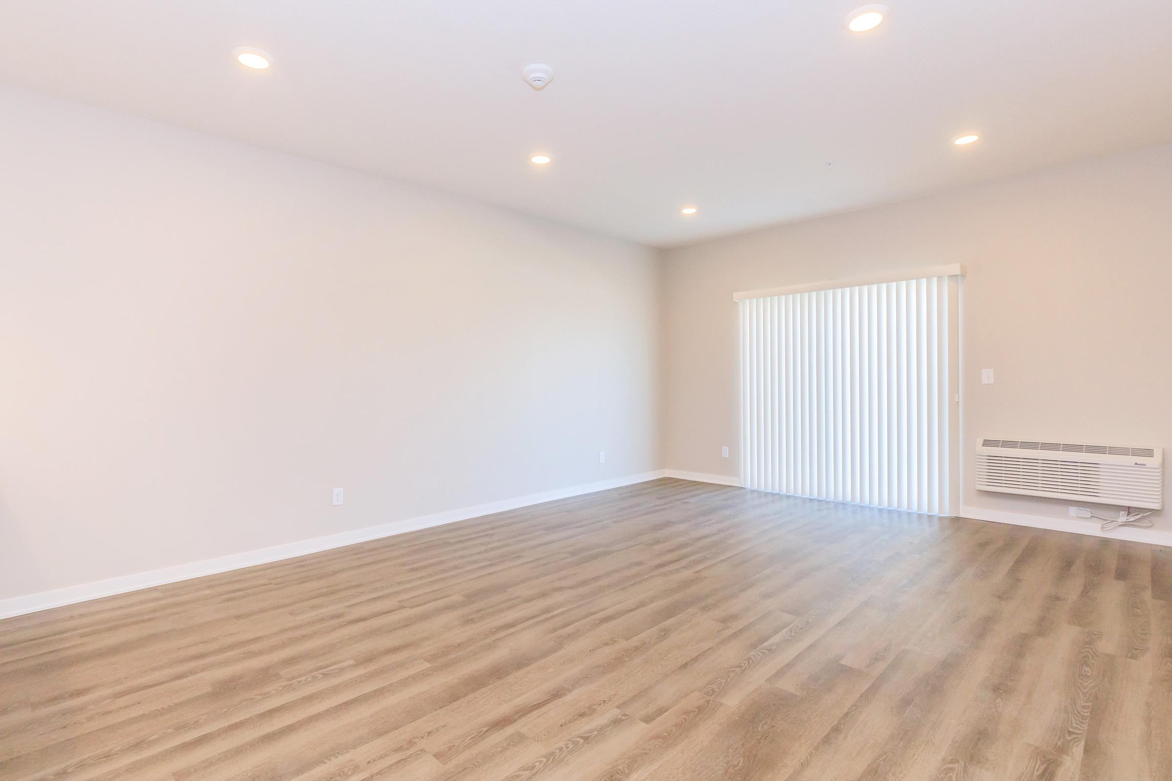 A spacious and empty living room with light-colored walls and wooden flooring. The room features a large window with vertical blinds, allowing natural light to enter. There is an air conditioning unit on one wall, creating a modern and minimalist atmosphere.