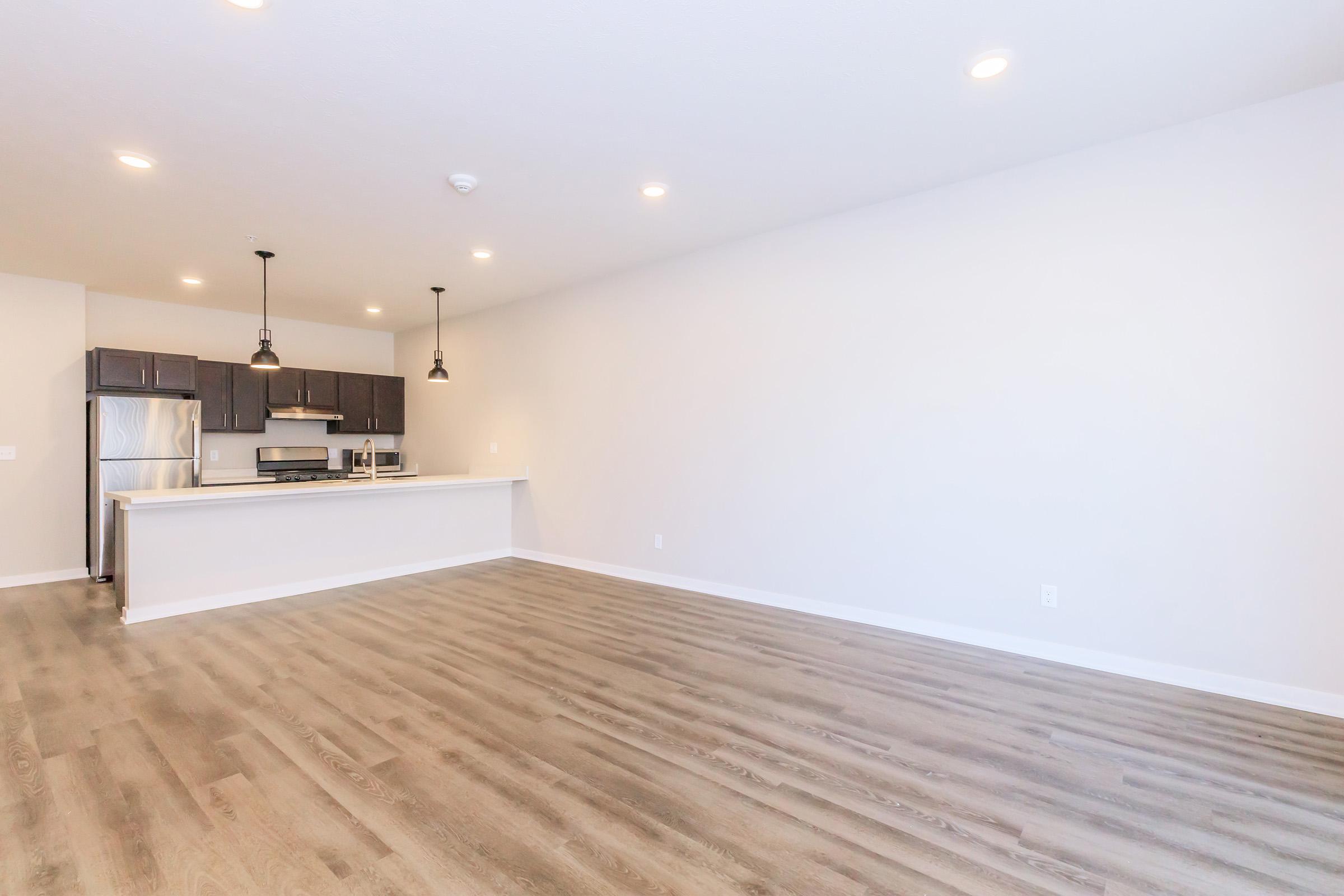 Spacious, empty living area featuring light-colored walls and hardwood flooring. A modern kitchen with dark cabinetry and stainless steel appliances is visible in the background, complemented by pendant lighting above the kitchen island. The room has ample natural light and a minimalistic design.