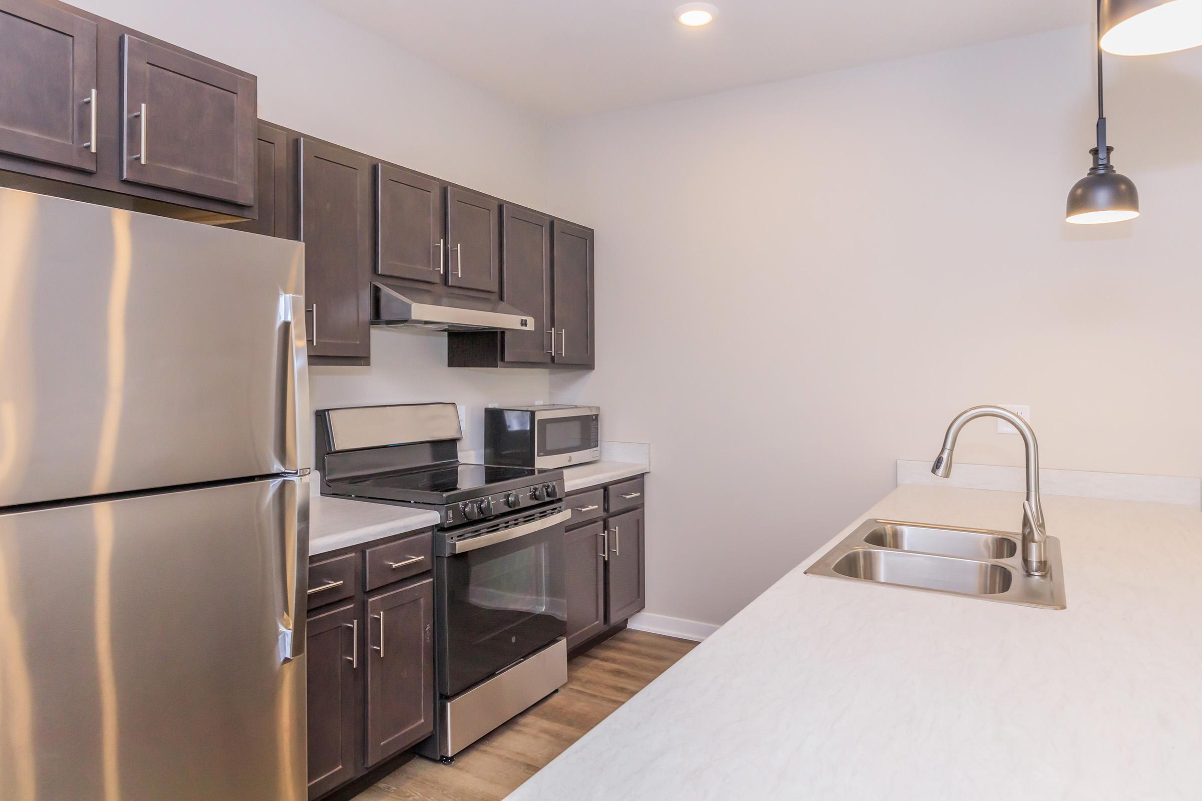 Modern kitchen featuring dark wood cabinetry, stainless steel appliances including a refrigerator and stove, and a sleek countertop with a built-in sink. The space is well-lit with overhead lighting, and there is a microwave and ample counter space for preparation. Neutral wall colors create a clean, inviting atmosphere.