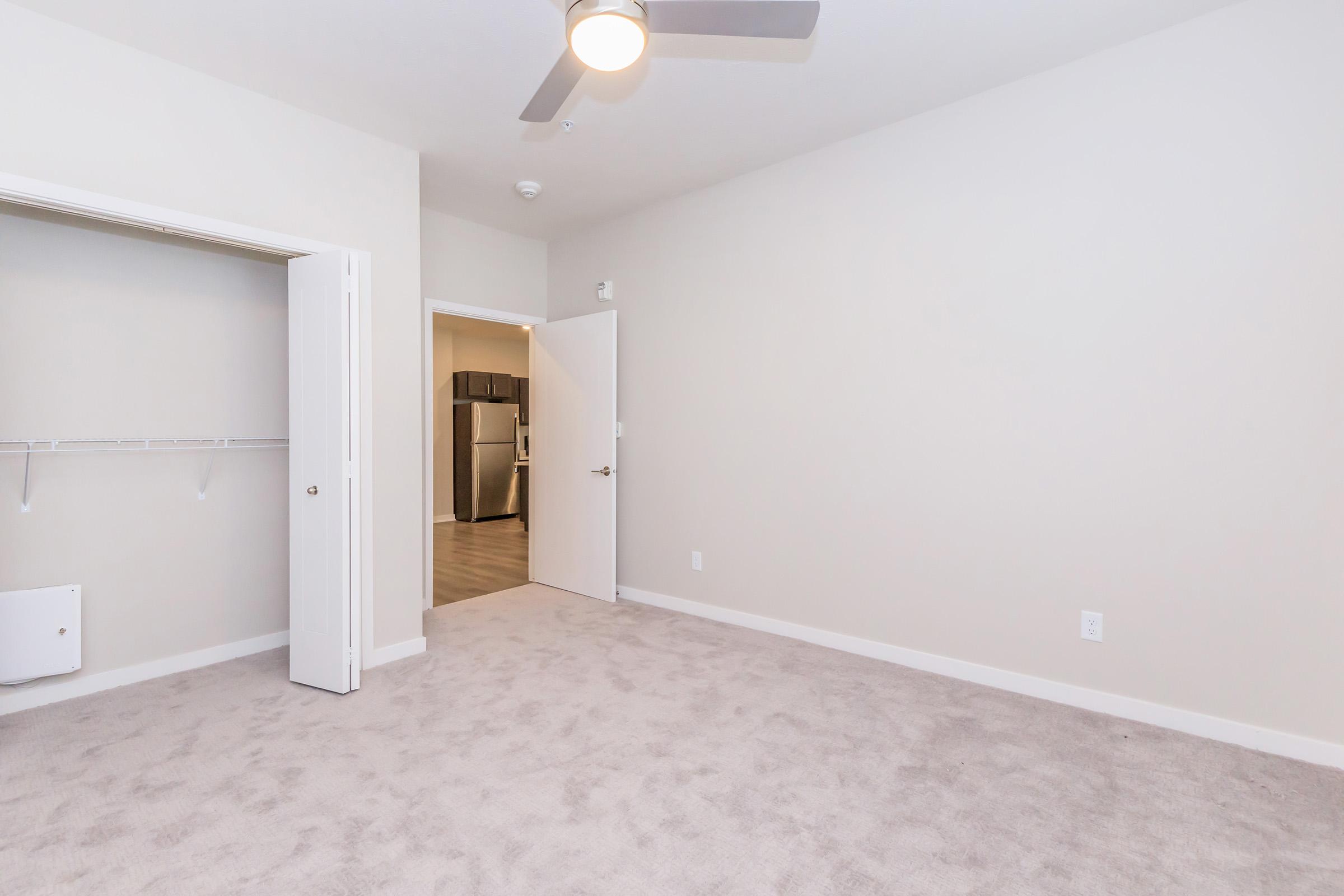 A spacious, light-filled room with light beige walls and plush carpet. An open closet is visible on the left, and a doorway leads to a kitchen area in the background. A ceiling fan is mounted above, adding functionality to the comfortable space. The overall ambiance is modern and inviting.