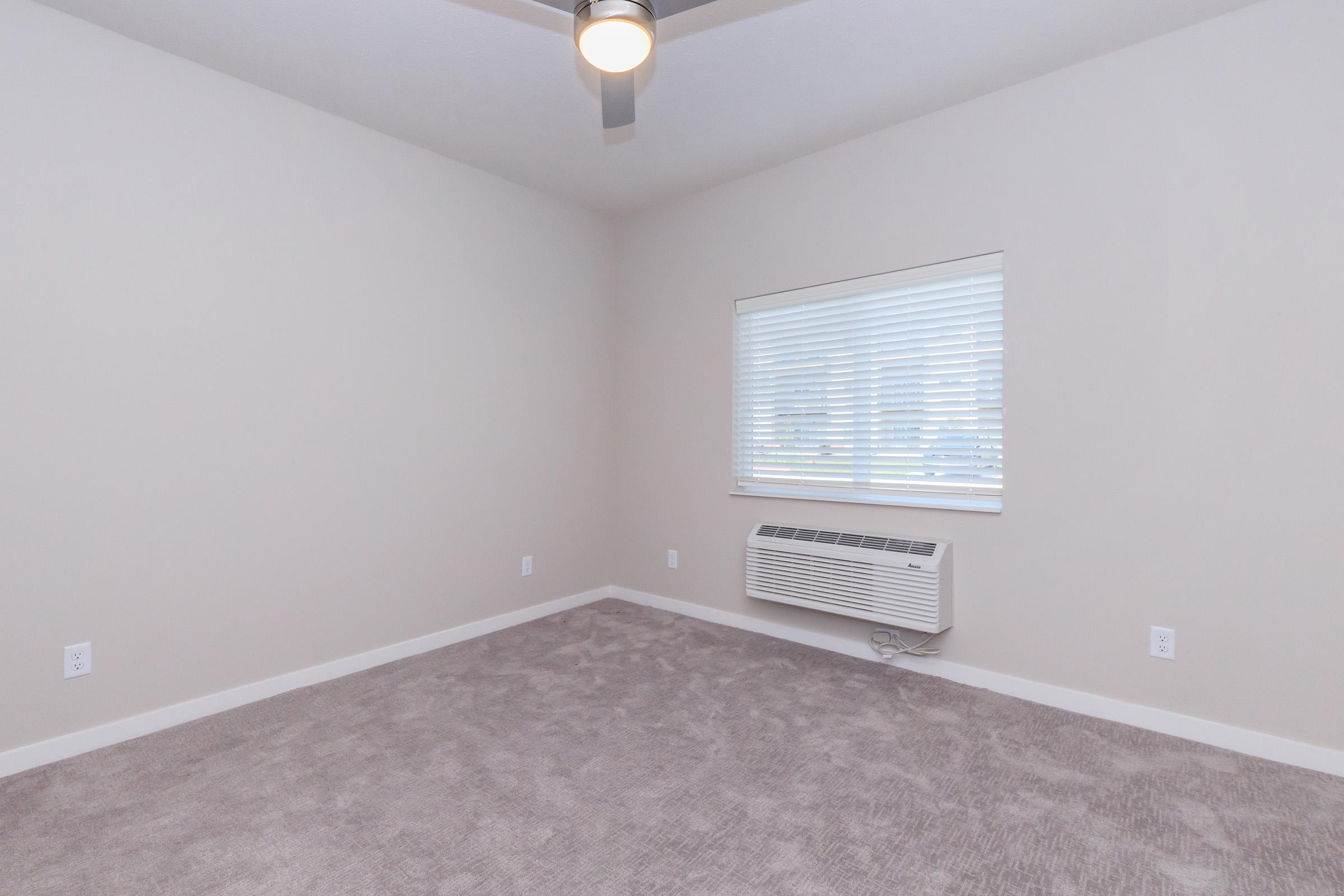 Empty room with light gray walls and a ceiling fan. The floor is covered in soft, light-colored carpet. A window with horizontal blinds lets in natural light, and an air conditioning unit is mounted beneath the window. The overall space appears bright and inviting.