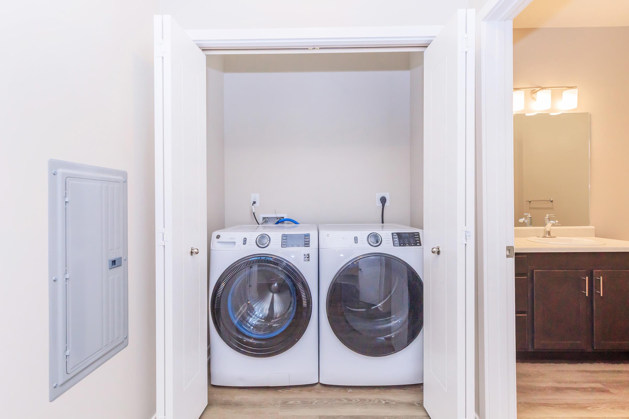 A clean, organized laundry room featuring a set of modern front-loading washing machines positioned side by side within white double doors. Light beige walls and wooden flooring create a bright and airy atmosphere. A glimpse of a bathroom vanity with a sink and mirror is visible in the background.