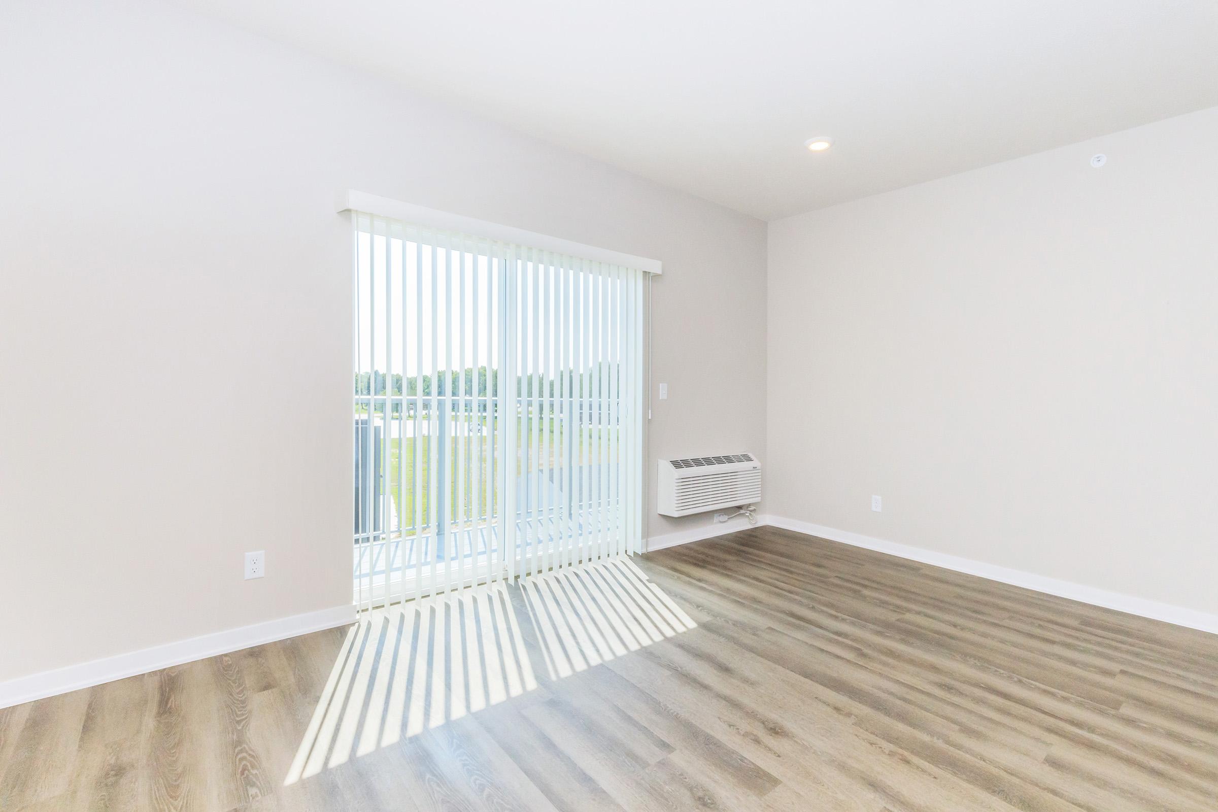 Empty room with light-colored walls and laminate flooring. There are vertical blinds covering a large sliding glass door that leads to a balcony. An air conditioning unit is mounted on the wall. Natural light casts striped shadows on the floor.