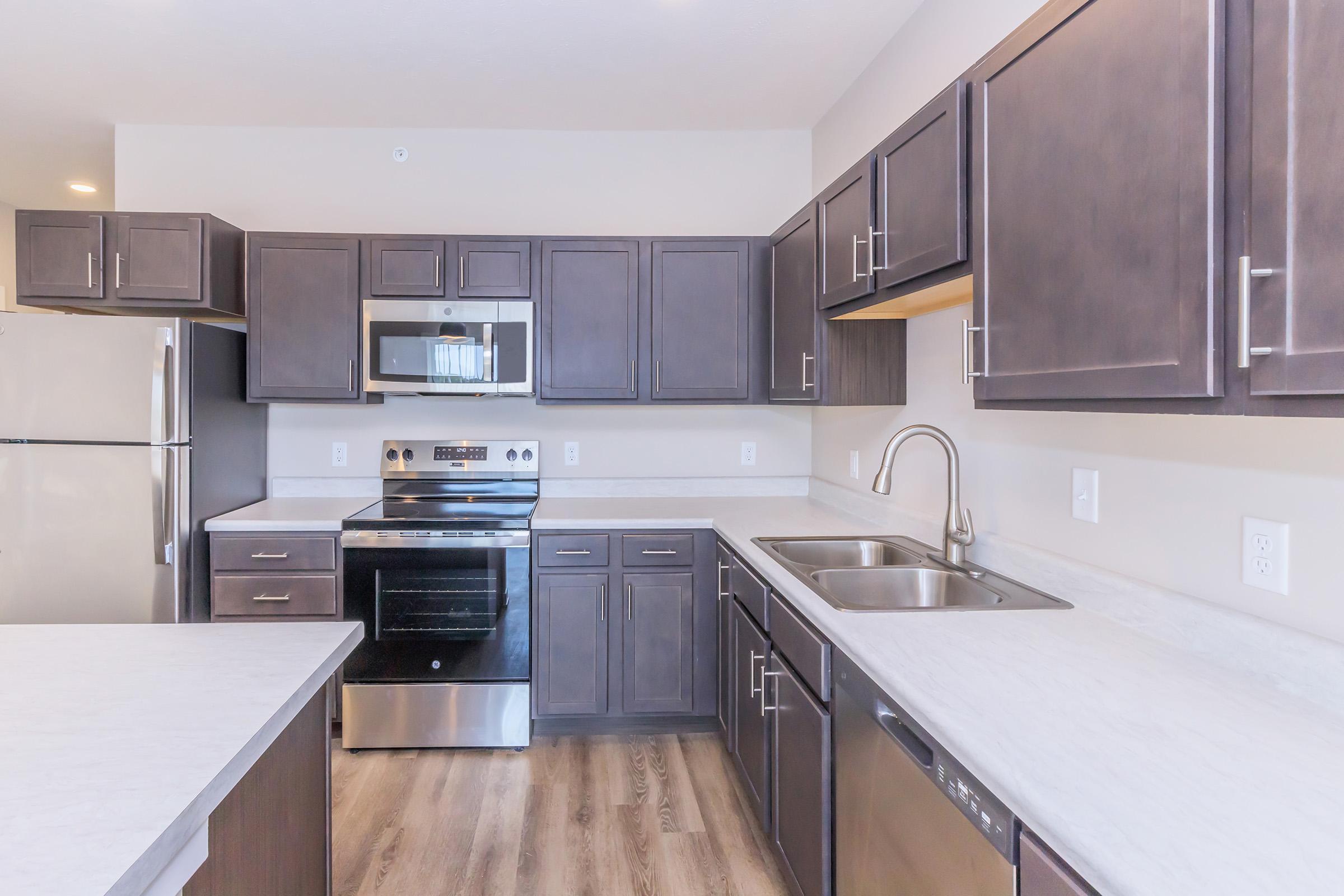 A modern kitchen featuring dark wood cabinets, stainless steel appliances including an oven, microwave, refrigerator, and dishwasher. The countertop is light-colored with a double sink and an open layout, providing ample workspace and storage. The flooring is wood-like, enhancing the contemporary design.