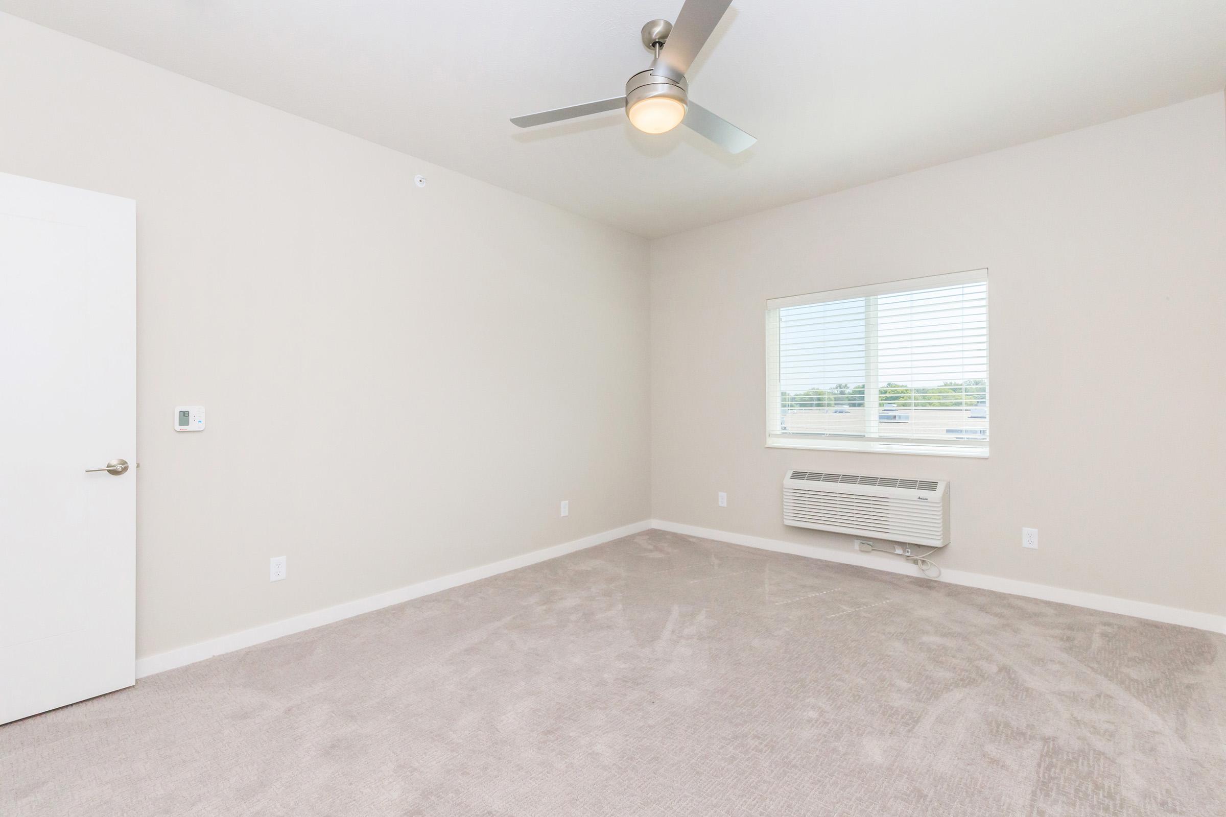 A bright, empty room featuring light-colored walls and carpet. There is a window with blinds allowing natural light, a ceiling fan, and an air conditioning unit on the wall. The door is closed and the space is uncluttered, giving a clean and open feel.