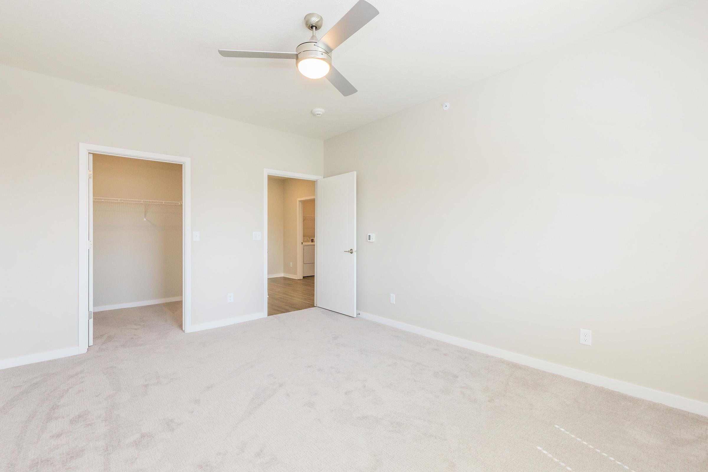 A spacious, empty bedroom featuring light beige walls, a ceiling fan, and soft carpet. A closet space is visible, along with an open door leading to a bathroom. The room is well-lit with natural light, creating a clean and airy atmosphere. There are no furnishings present.