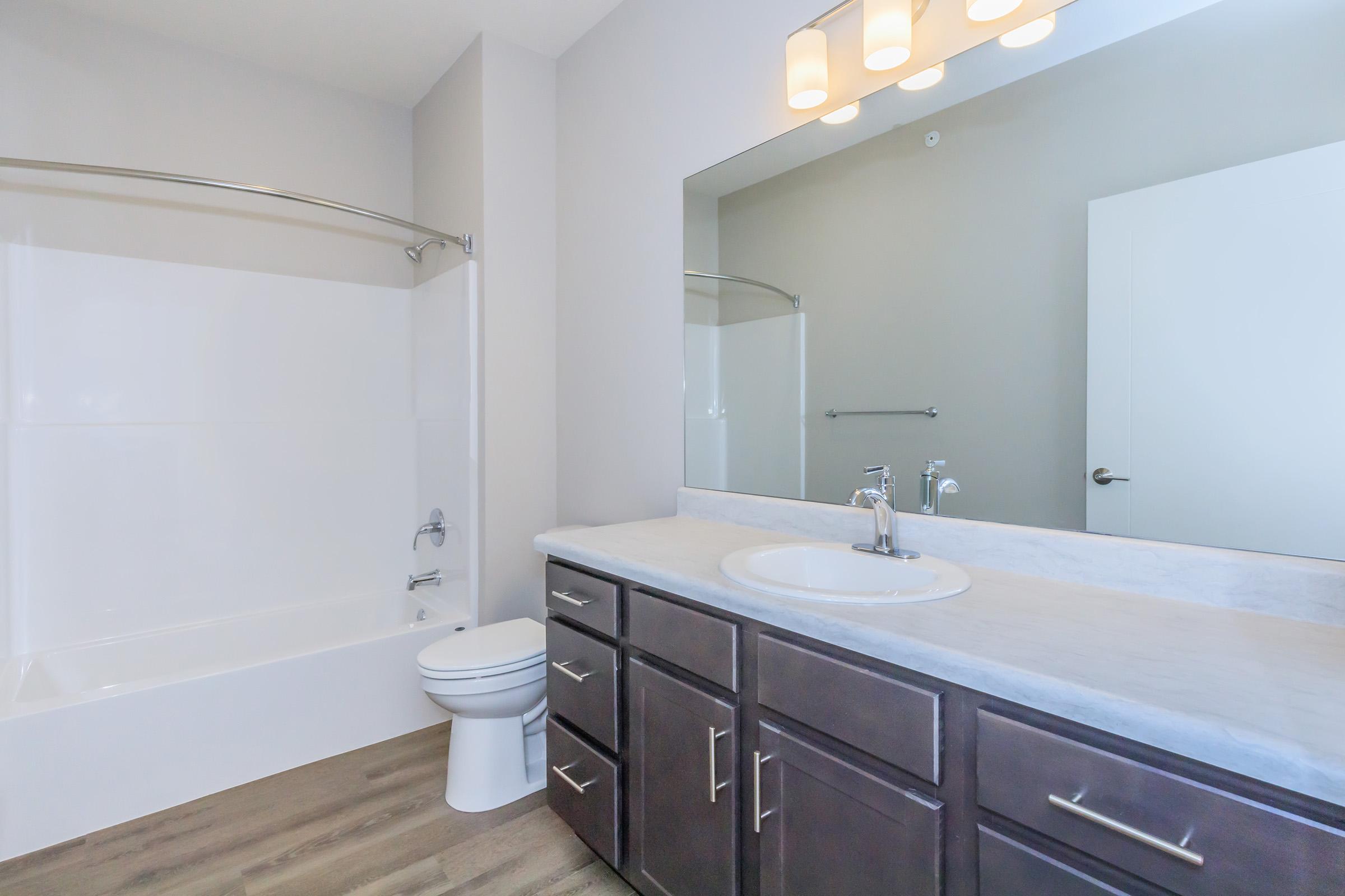 A modern bathroom featuring a tub and shower combination, a white countertop with a circular sink, and a large mirror above. The cabinetry is dark wood, and there is a white toilet. Soft lighting from overhead fixtures creates a bright atmosphere, complemented by neutral wall colors and wood-like flooring.