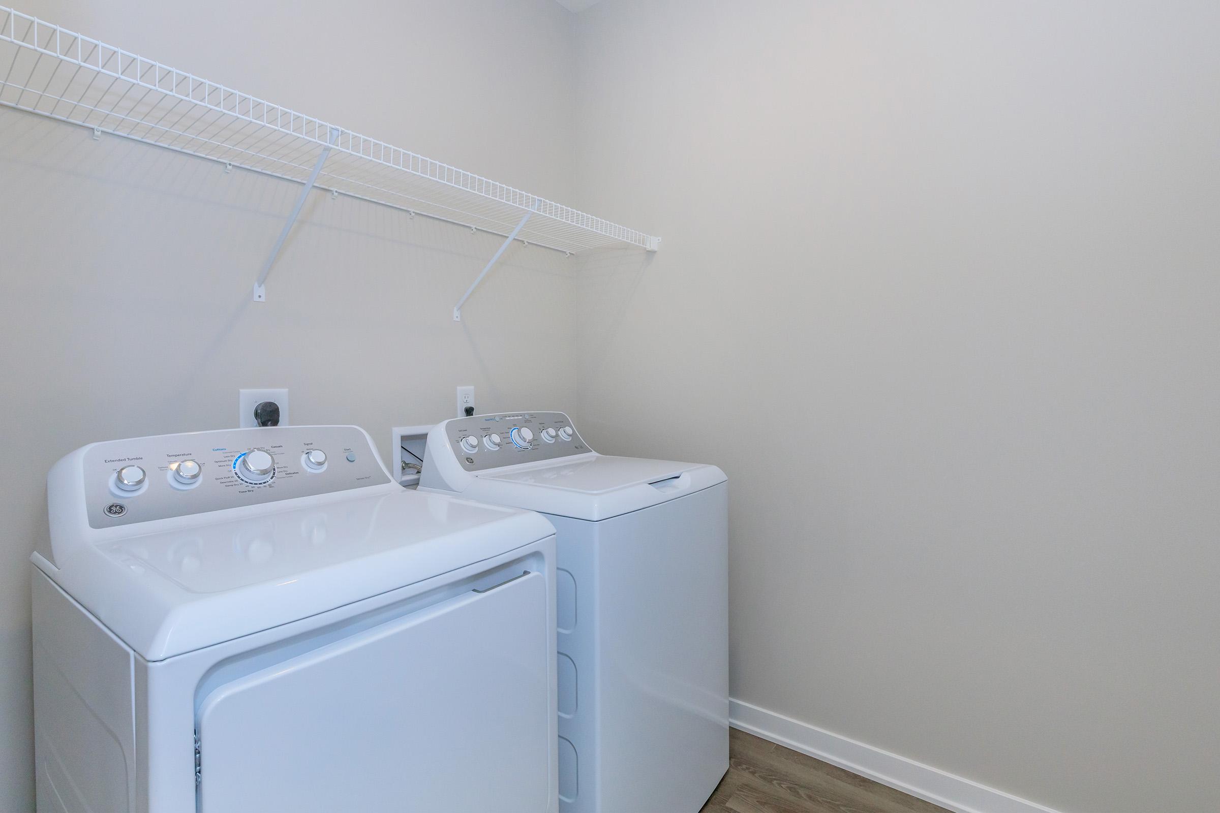 A clean laundry room featuring two white appliances: a top-loading washing machine and a front-loading dryer. Both machines are placed side by side against a light-colored wall. Above them, a wire shelf provides additional storage space, and the floor is a light wood finish.