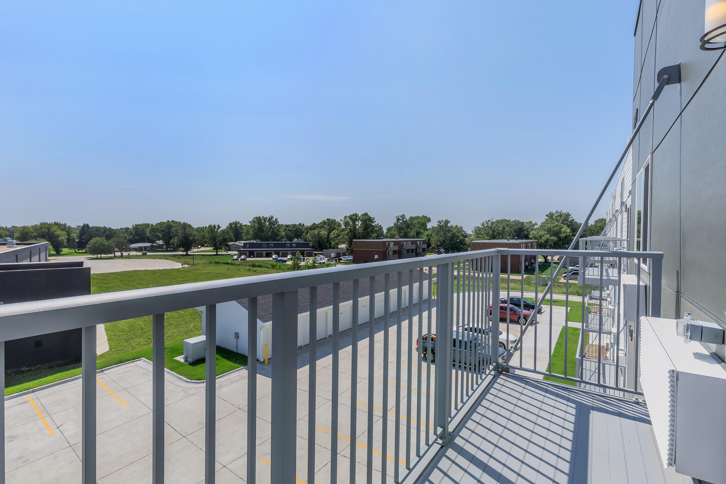 View from a balcony overlooking a parking lot and green landscape. The sky is clear with few clouds, and there are several buildings in the background, including low-rise apartments. The railing is a light gray color, providing a modern touch to the outdoor space.