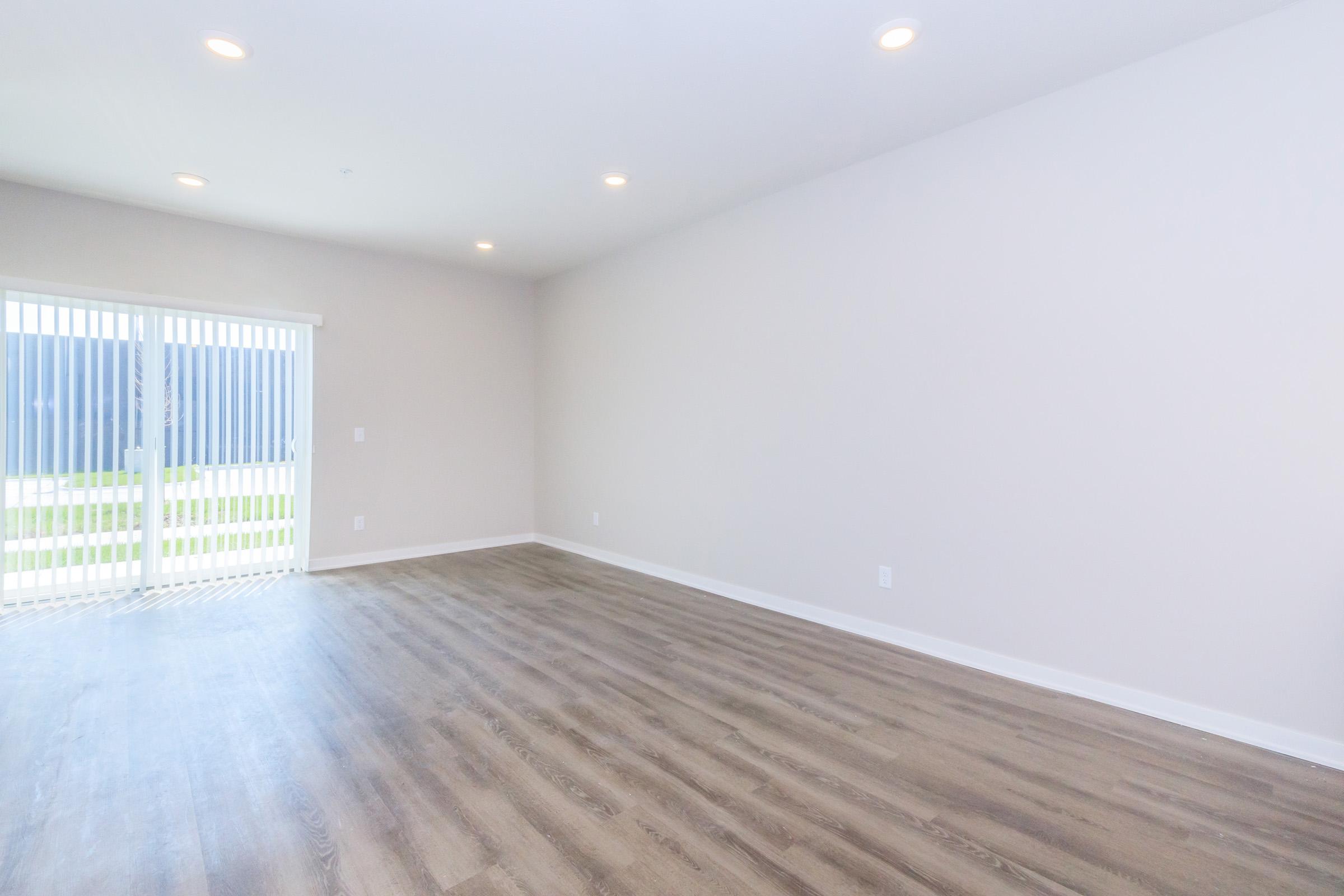 A spacious, empty room with light-colored walls and wooden flooring. It features large sliding glass doors that open to a grassy outdoor area, allowing natural light to fill the space. The ceiling is equipped with recessed lighting, enhancing the bright and airy atmosphere.