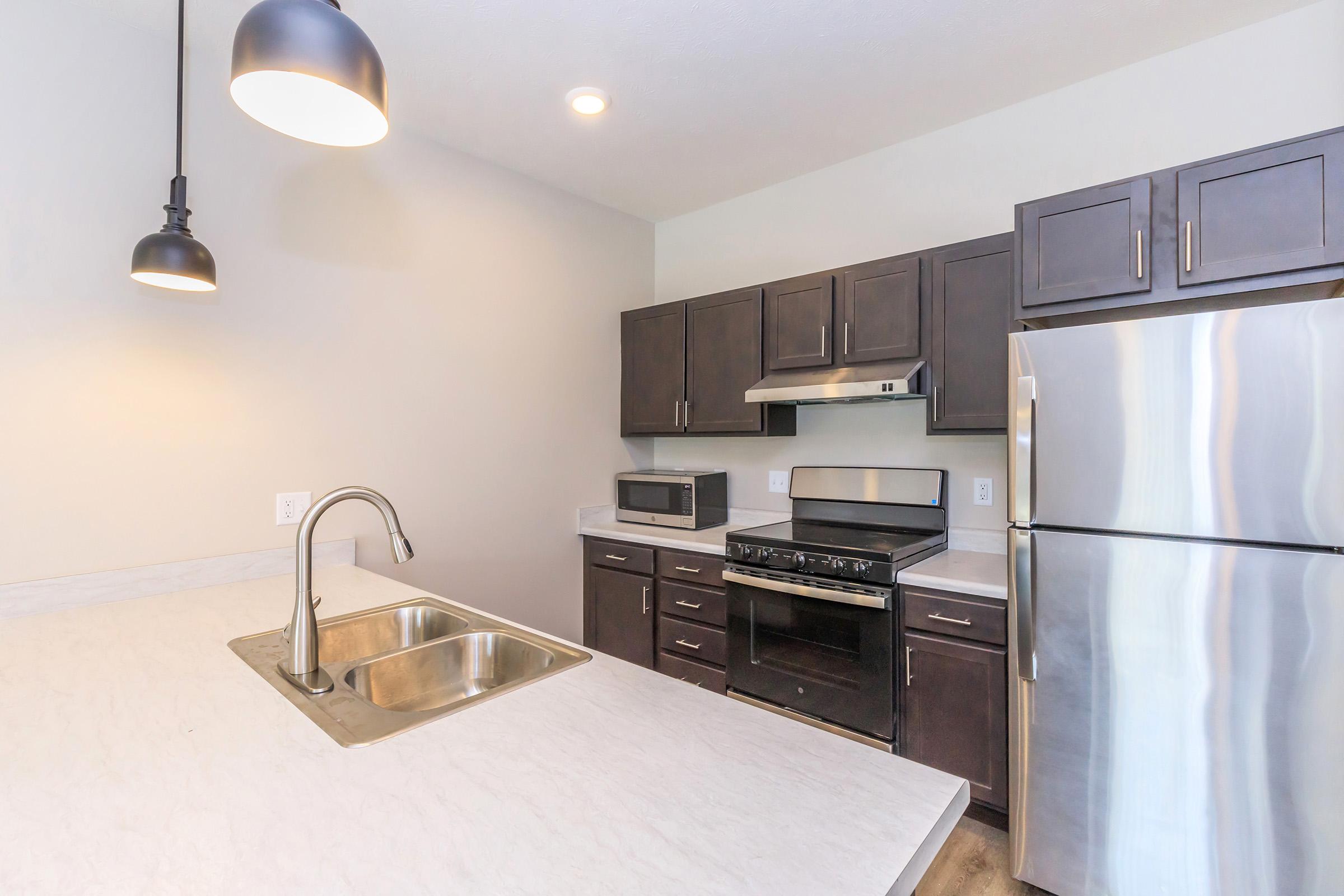 Modern kitchen featuring a light-colored countertop, stainless steel sink, dark wooden cabinets, a silver refrigerator, and a black oven with a microwave above it. Ceiling lights provide illumination, and the walls are painted in a light neutral shade, creating an inviting and functional cooking space.