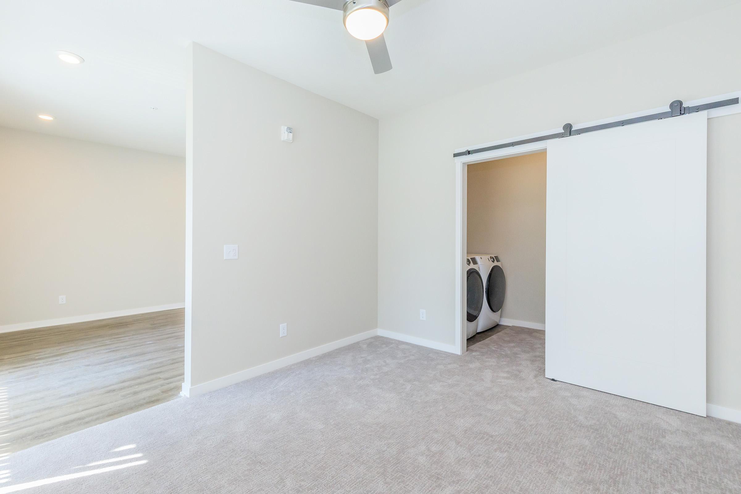 Interior view of a small, clean, and well-lit room with light-colored walls and plush carpet. There is a ceiling fan and a sliding door that leads to a closet area, which contains a washer and dryer. The room connects to a second, slightly larger area with natural light.