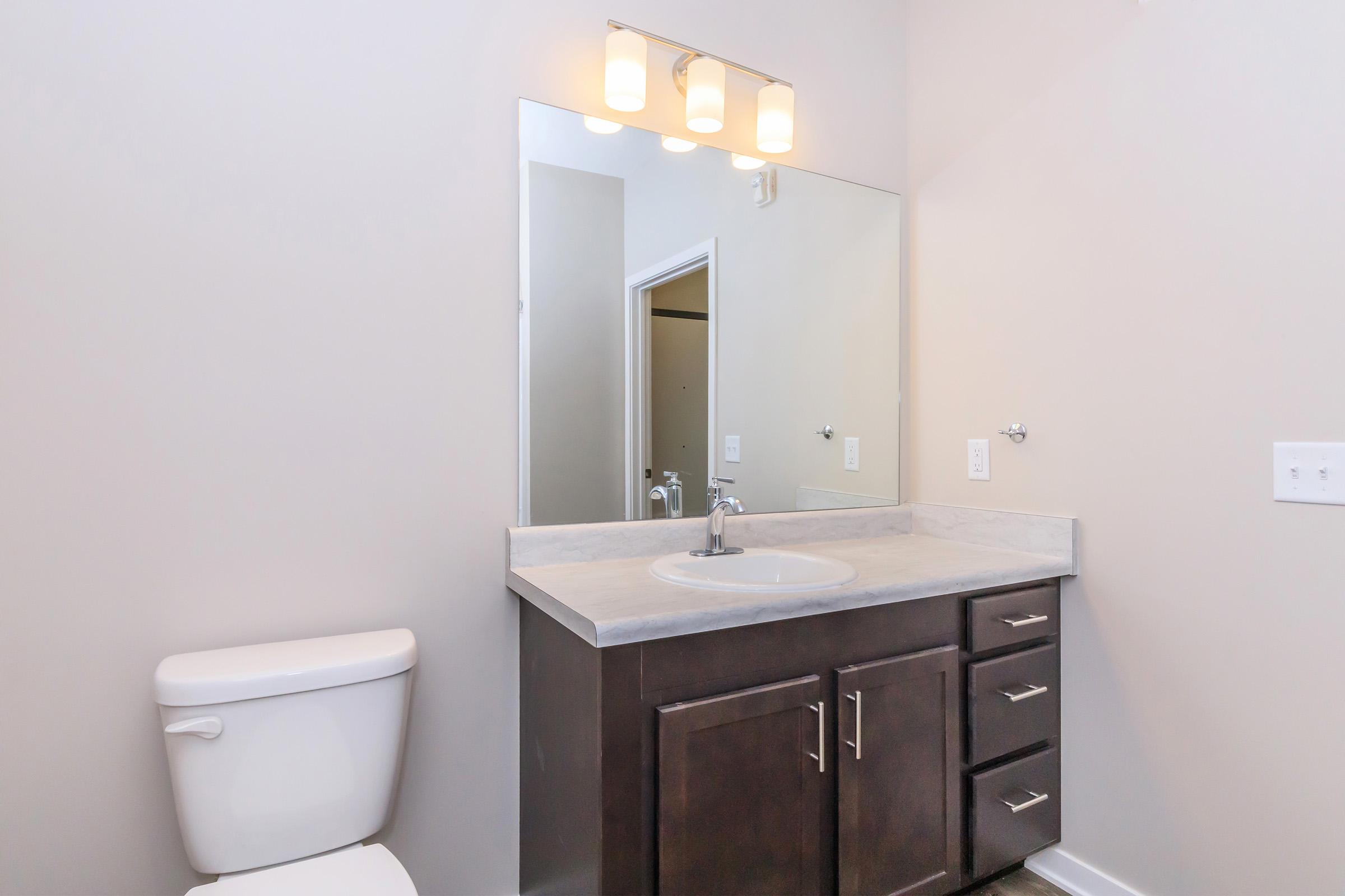 A modern bathroom featuring a single sink vanity with a gray countertop, dark wooden cabinets, a large mirror above the sink, a white toilet, and neutral-colored walls. The space is well-lit with overhead lighting and has a clean, minimalist design.