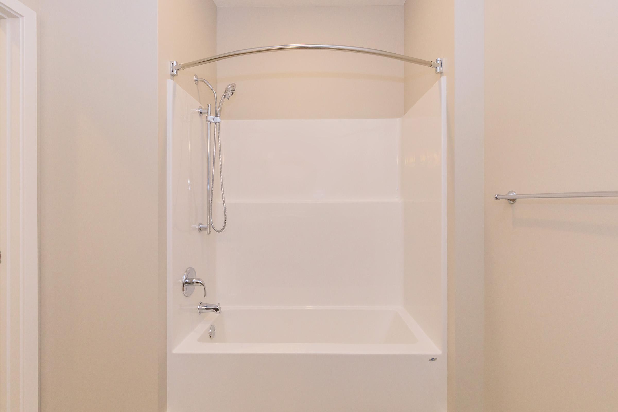 A clean, modern bathroom with a white shower and bathtub combination. The shower features a chrome showerhead and a curved glass shower rod. The walls are painted a light neutral color, and there is a towel bar on one side, providing a minimalist and functional design.