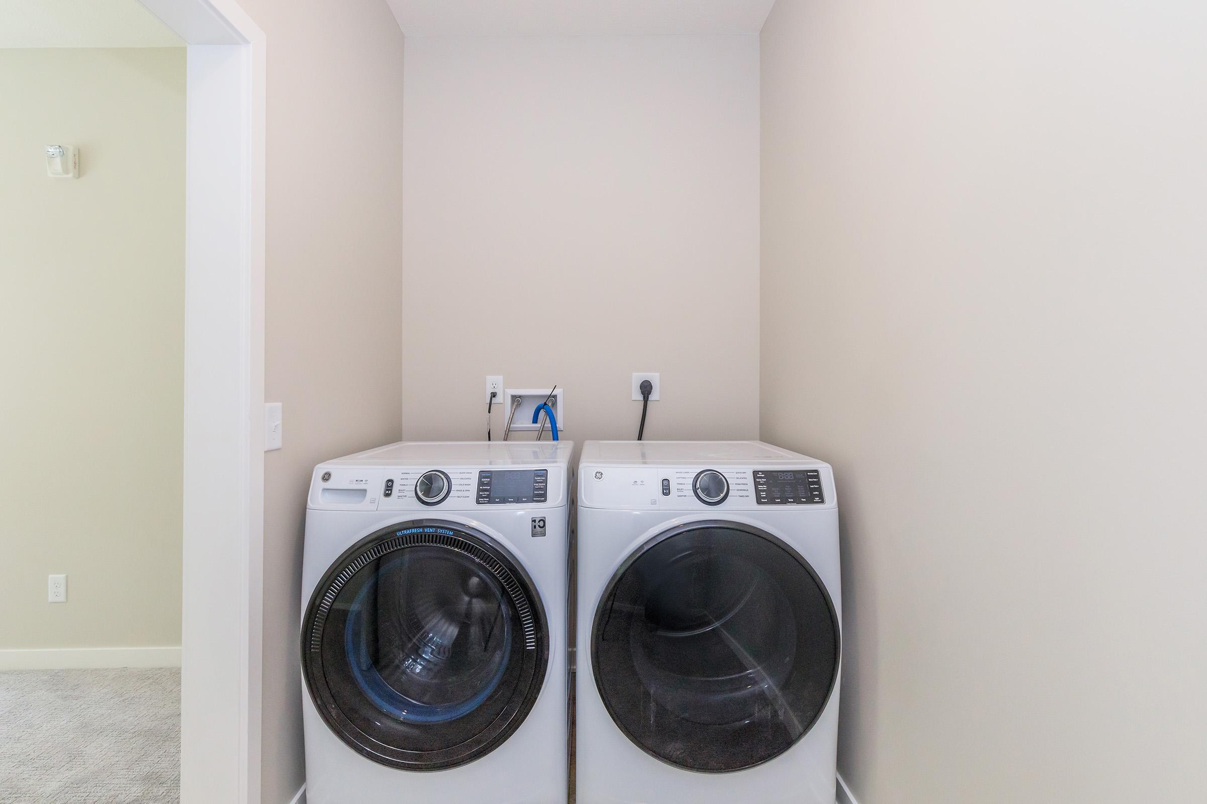 A modern laundry room featuring two white front-loading washing machines side by side against a light beige wall. The machines have control panels facing forward, and there are utility connections visible behind them. The floor is carpeted in a light color.