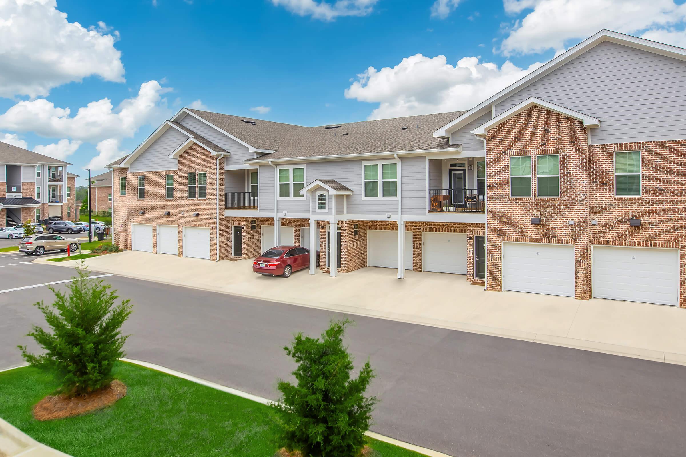 A residential apartment building with brick and light gray exterior. The structure features multiple garages and balconies. A red car is parked in front of one garage. Lush green grass and small trees line the driveway, and the sky is bright with fluffy white clouds.