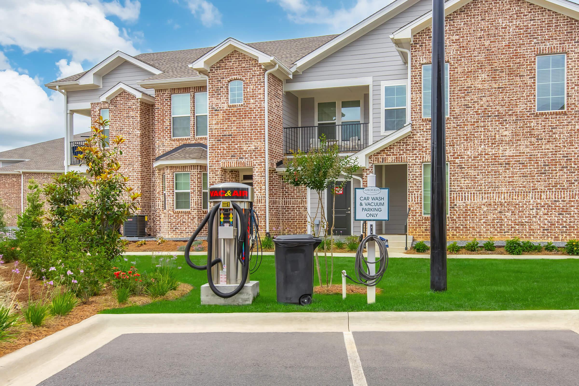 Air pump station for vehicles located in front of a brick apartment building. The area includes a sign indicating "Car Wash Parking Only," a trash can, and hoses connected to the pump, surrounded by well-maintained grass and landscaping. Bright sky with scattered clouds in the background.