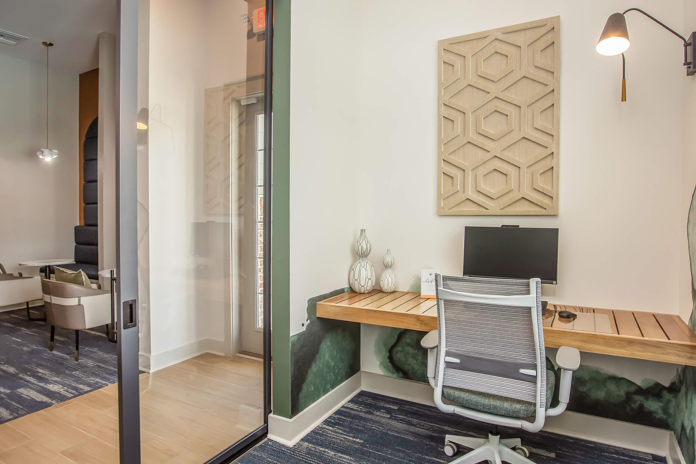 A modern office space featuring a wooden desk with a computer, a comfortable office chair, and decorative wall art. The room has a glass door leading to a lounge area and is decorated with soothing colors and textures, creating a professional and inviting atmosphere.