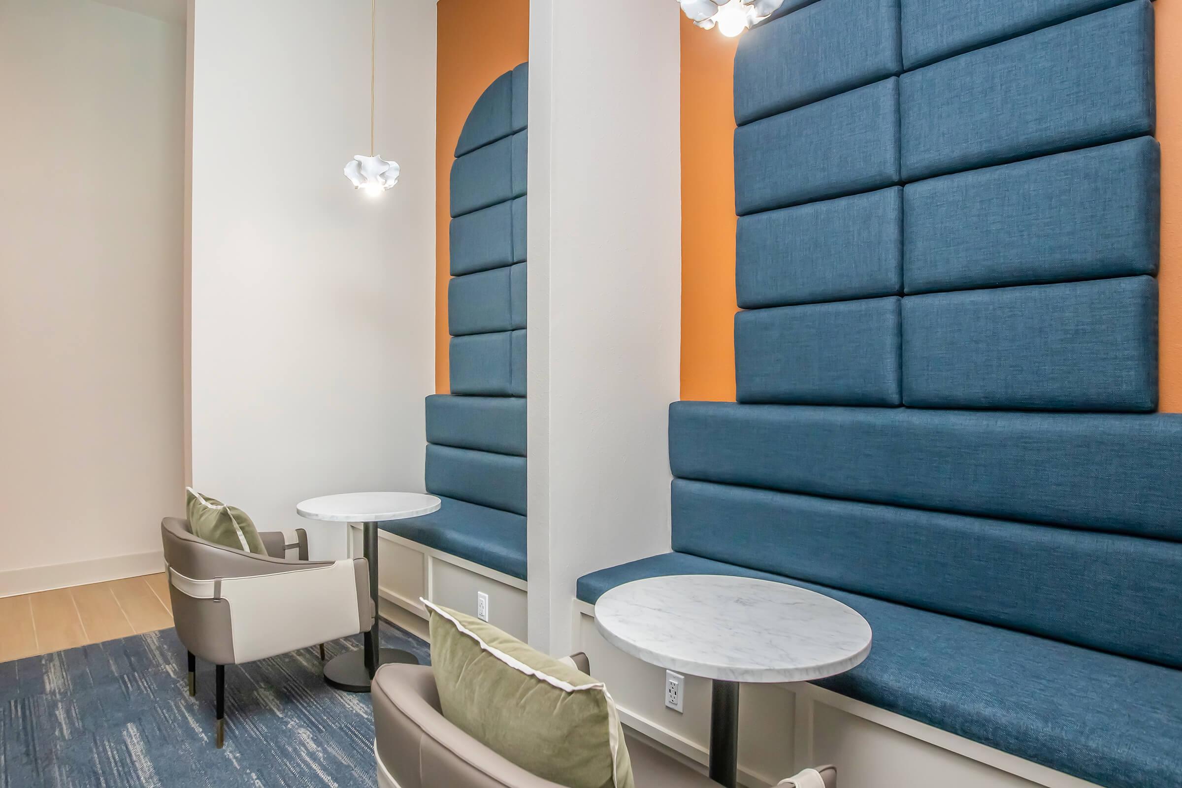 Modern lounge area featuring two blue upholstered benches against a white wall, each accompanied by a small round marble table. Gentle lighting from decorative fixtures above creates a welcoming atmosphere. The space has a minimalist design with a touch of warmth from an orange accent wall.
