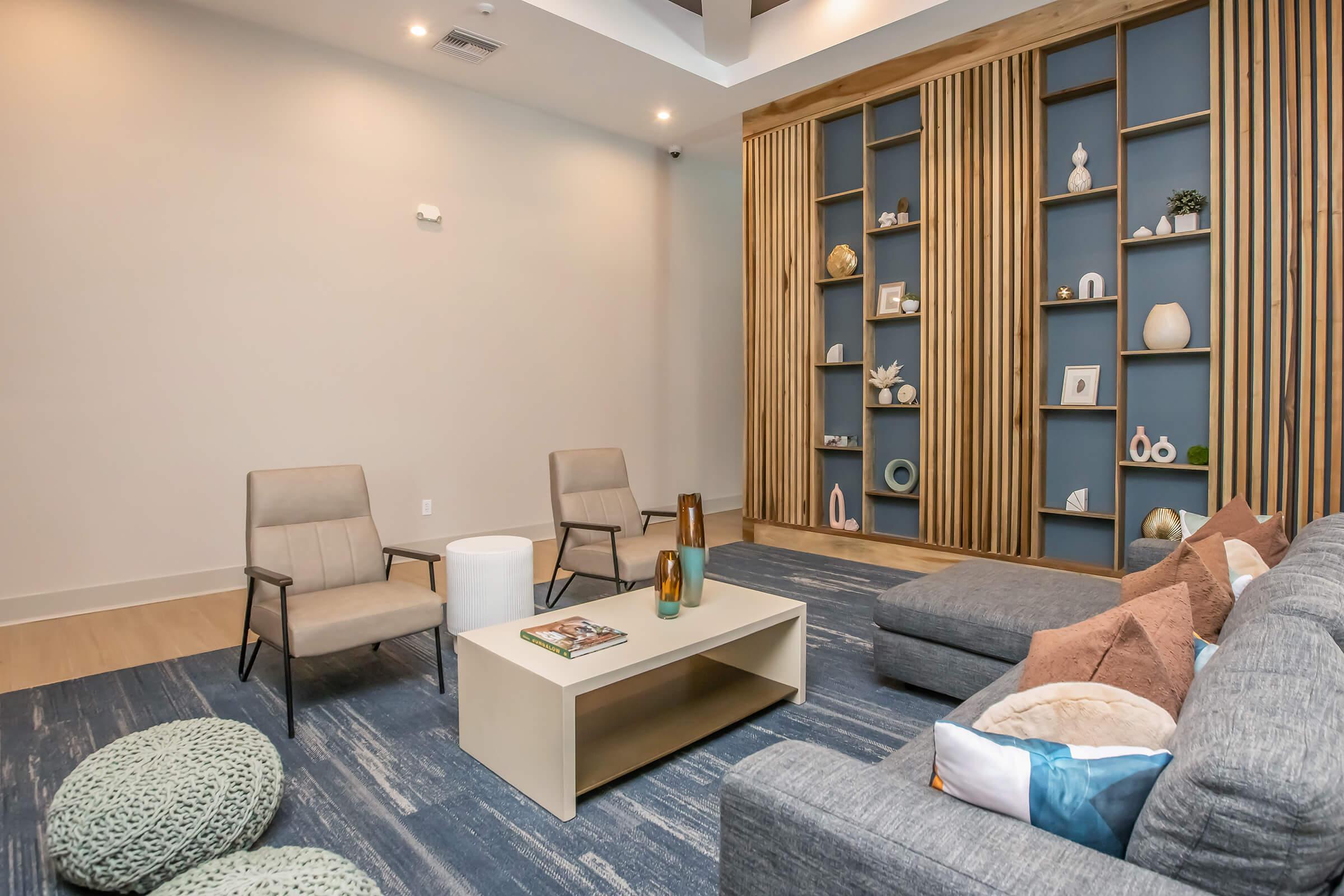 A modern living space featuring two gray armchairs and a light-colored coffee table in the center. A stylish bookshelf with decorative items in various colors and textures is mounted on the wall. The floor is covered with a blue area rug, and there are plush cushions scattered around.
