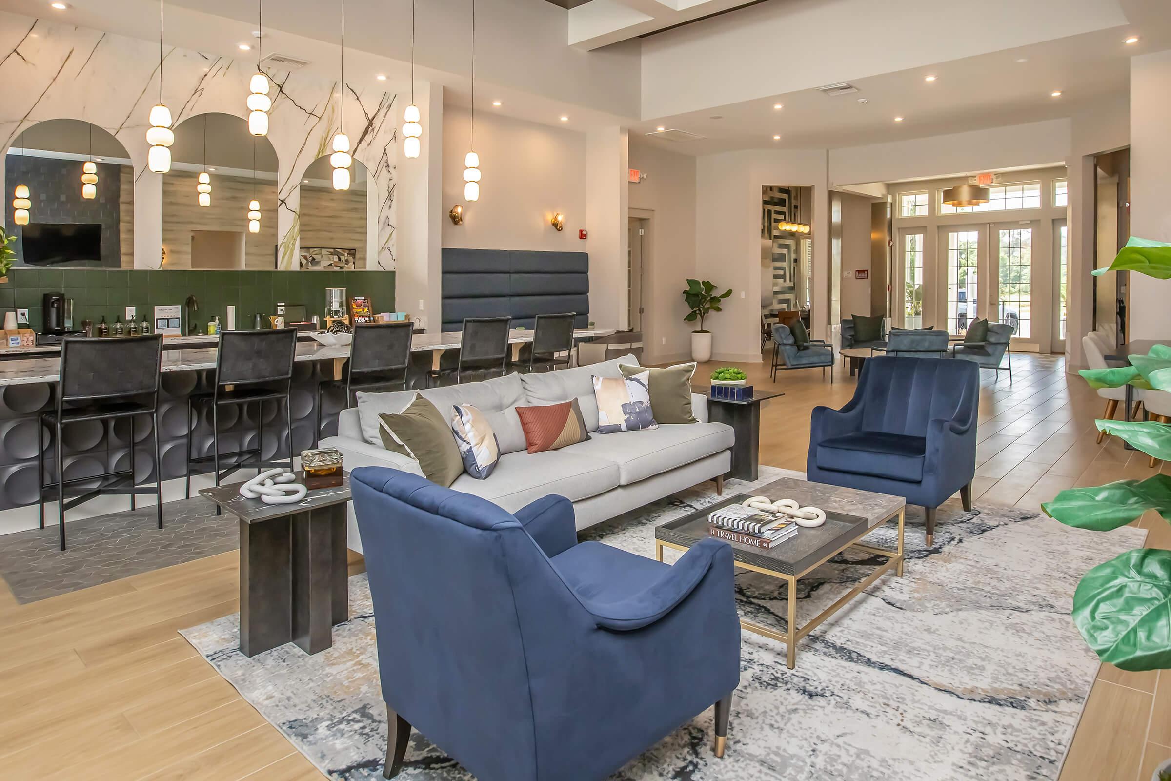A modern lounge area featuring a light gray sofa with colorful cushions, two dark blue armchairs, and a coffee table on a patterned rug. There is a bar area with high stools in the background, plants, and large windows allowing natural light into the space. Elegant light fixtures hang from the ceiling, adding a warm ambiance.