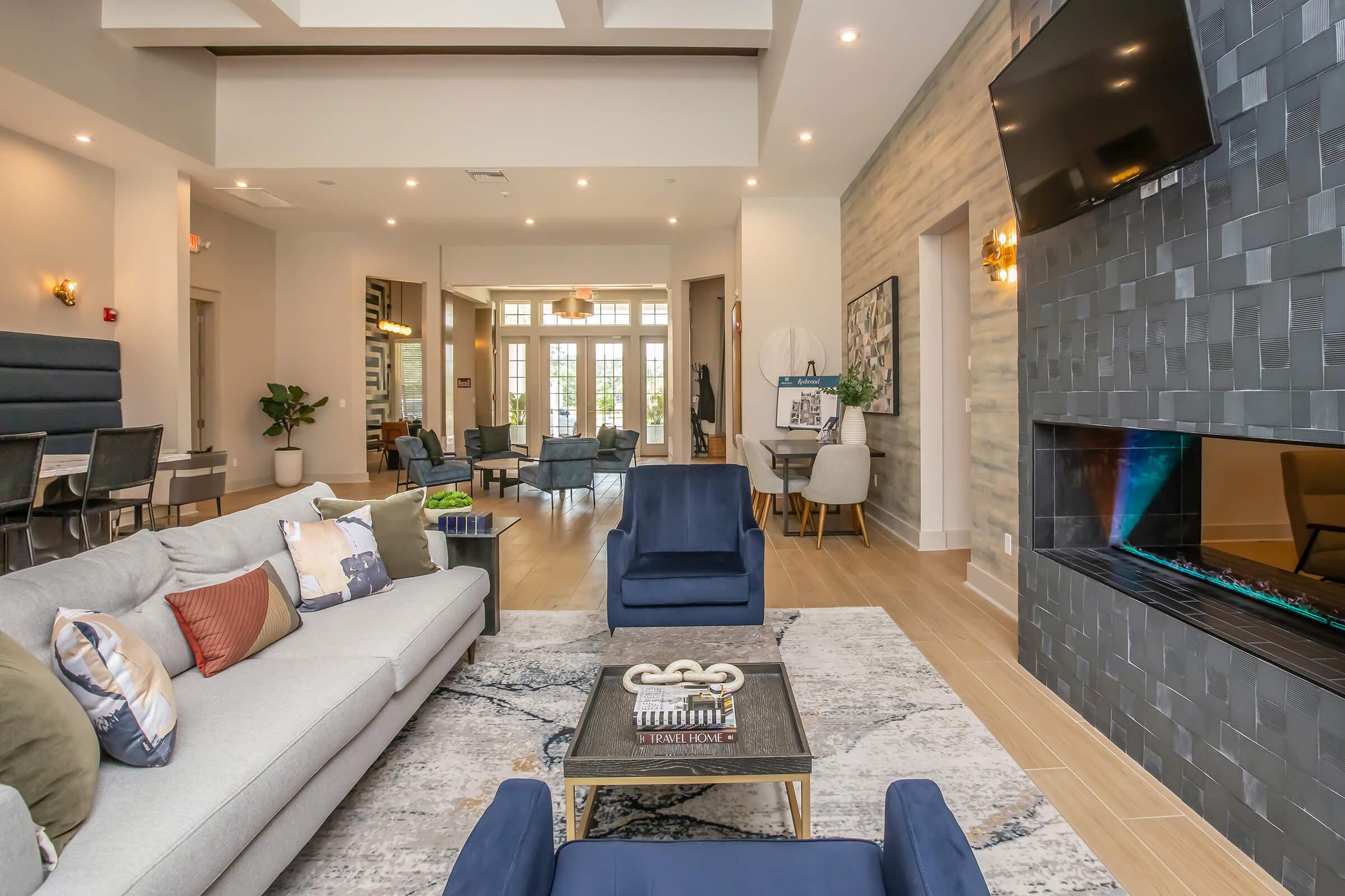 Modern lounge area with a light gray sectional sofa adorned with decorative pillows, a glass coffee table on a patterned rug, and a sleek fireplace. The space features contemporary furniture, ample natural light, and stylish decor, creating a welcoming atmosphere. A flat-screen TV is mounted on a feature wall.