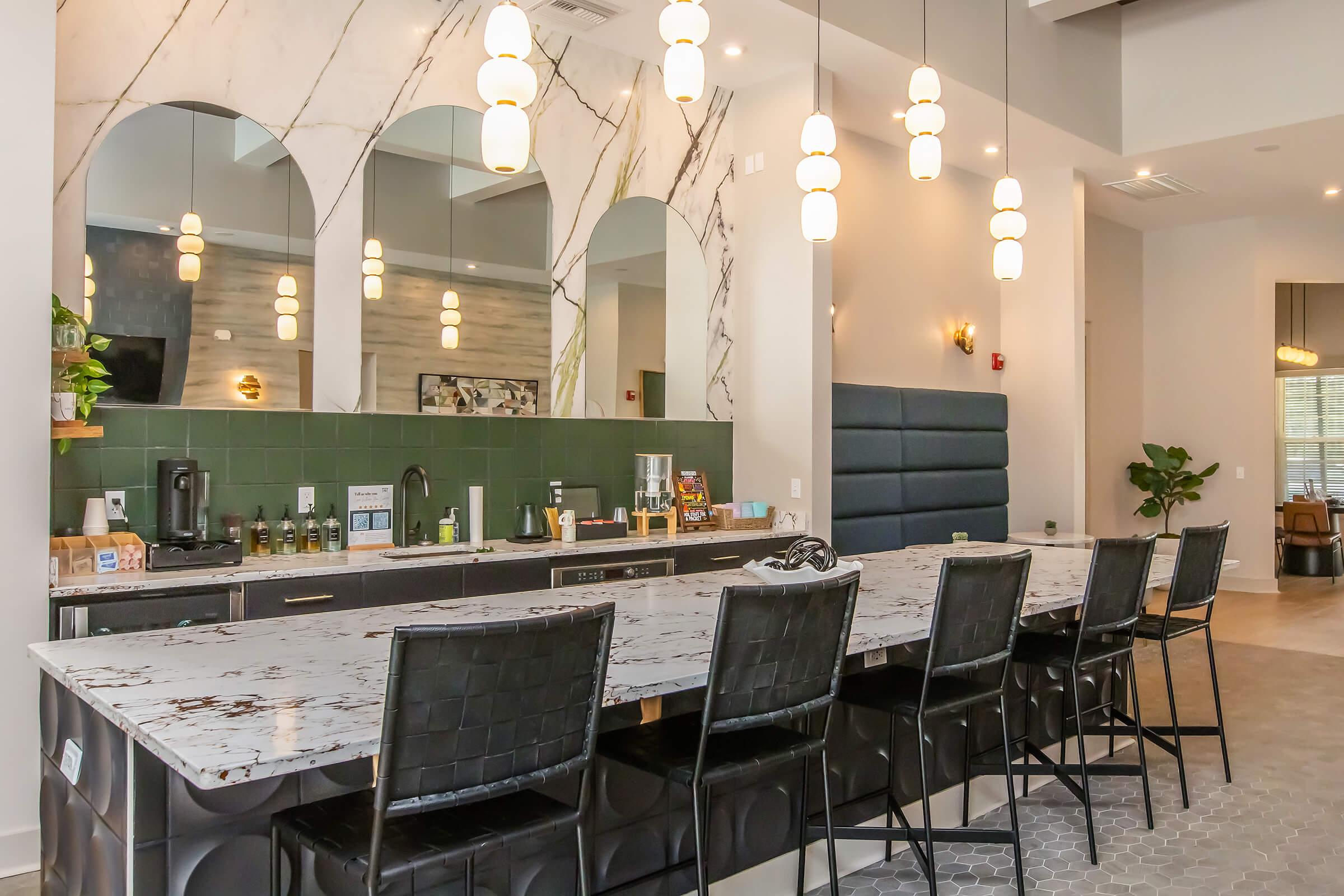 Modern kitchen with a large marble countertop and six black bar stools. Features stylish lighting fixtures, a green backsplash, and large mirrors above the countertop. The area is well-lit, with a plant in the corner and various kitchen appliances visible on the countertop.
