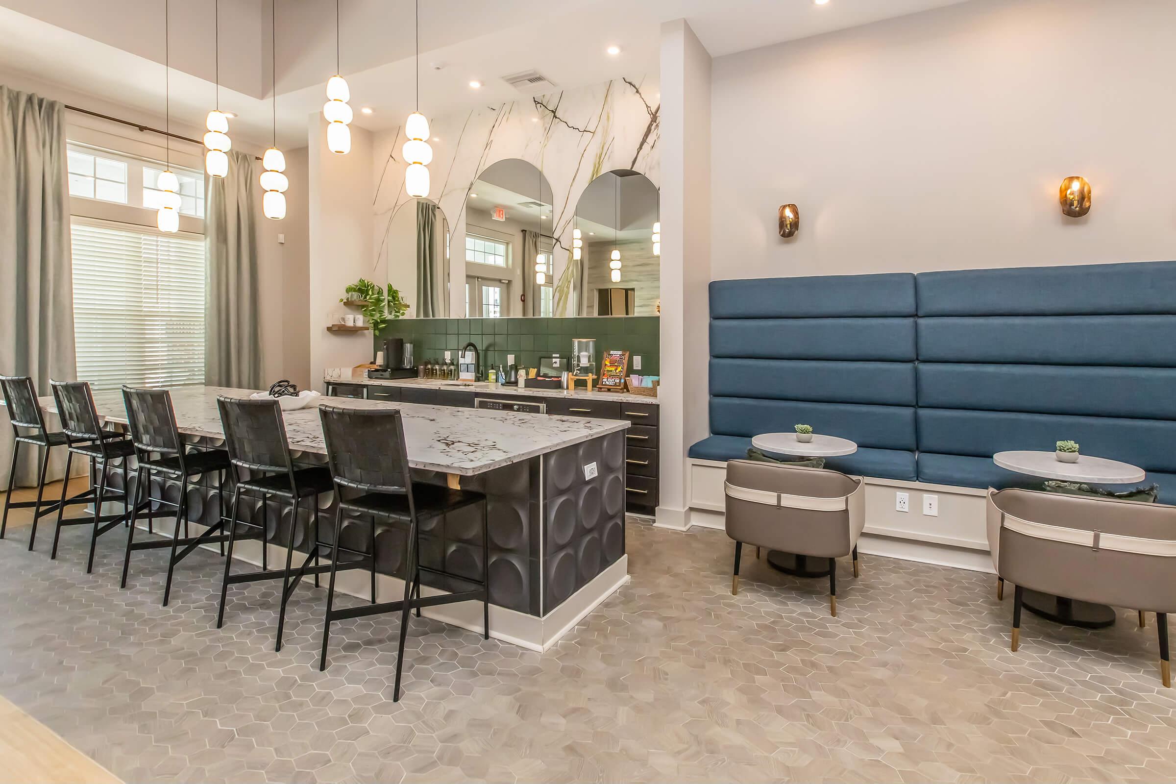 A modern interior of a stylish lounge featuring a long bar with high stools, a marble countertop, and two circular tables with low chairs. Large windows are draped with curtains, and decorative lighting adds ambiance. A mirrored wall reflects the space, enhancing the contemporary design.