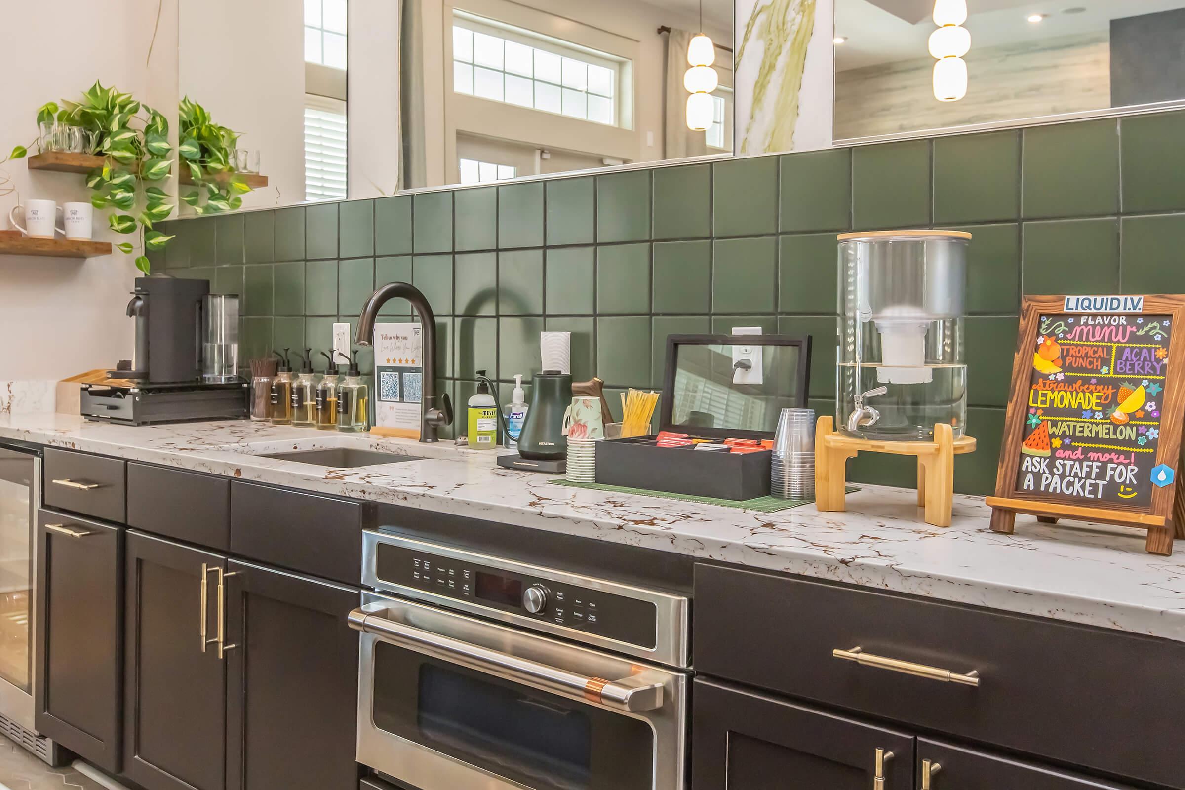 Modern kitchen with dark cabinetry and a marble countertop featuring a beverage station. Display includes a water cooler, condiments, cups, and a colorful sign with drink options. Green tiled backsplash adds a pop of color, complemented by natural light and indoor plants.