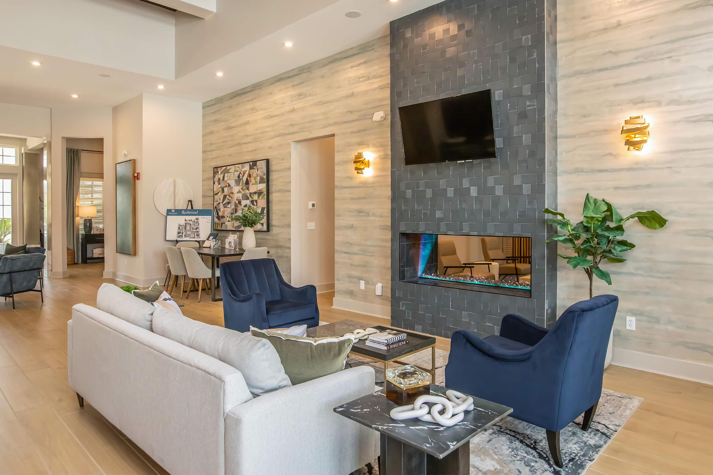 A modern living room featuring a light-colored sofa and two navy blue armchairs. The wall has a decorative stone finish and a mounted TV above a sleek linear fireplace. A coffee table with decorative items sits in front of the sofa, and a potted plant adds a touch of greenery to the space. Natural light brightens the room.