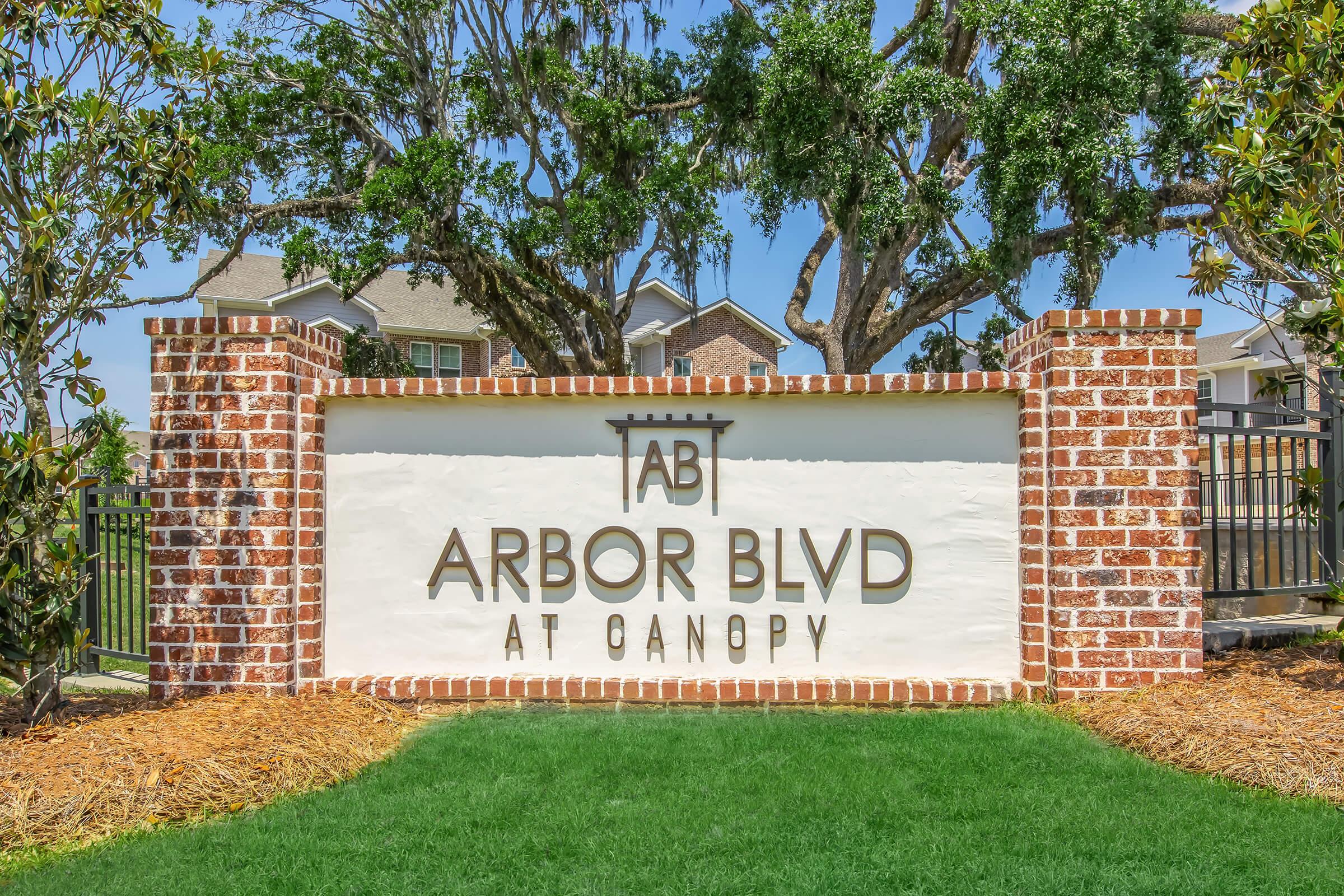 A sign for "Arbor Blvd at Canopy," featuring a modern design with brown lettering on a white background, bordered by brick pillars. The background includes green grass and large trees, indicating a landscaped area in a residential or community setting.