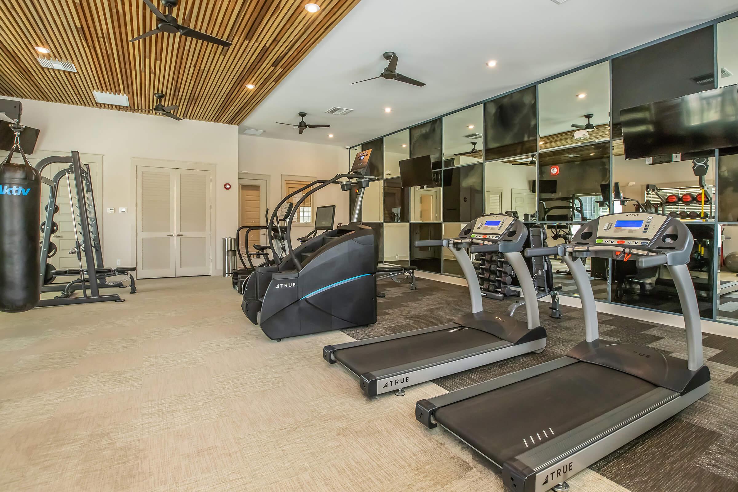 A modern fitness center featuring multiple exercise machines, including treadmills, an elliptical machine, and a punching bag. The space has a stylish wooden ceiling, mirrors on one wall, and various workout equipment, creating a bright and inviting atmosphere for physical activities.