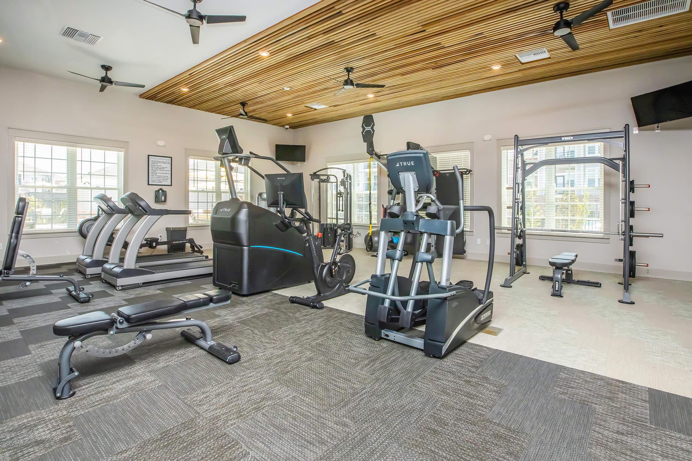Modern gym interior featuring various fitness machines, including treadmills, an elliptical, strength training equipment, and benches. The space has large windows for natural light, a wooden ceiling, and overhead fans, creating a bright and inviting atmosphere for workouts.