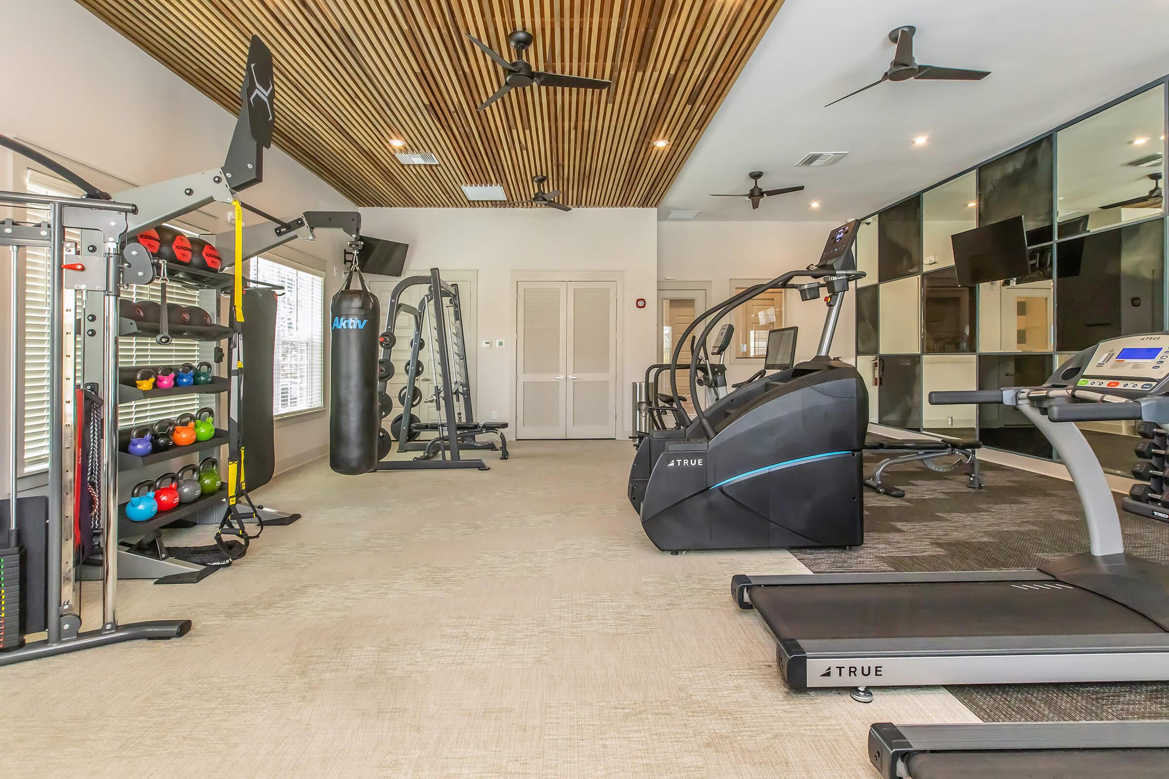 A modern gym interior featuring a variety of fitness equipment including treadmills, an elliptical machine, a punching bag, weight racks with colorful dumbbells, and exercise machines. The room has a wood-paneled ceiling and ample natural light, creating an inviting workout environment.