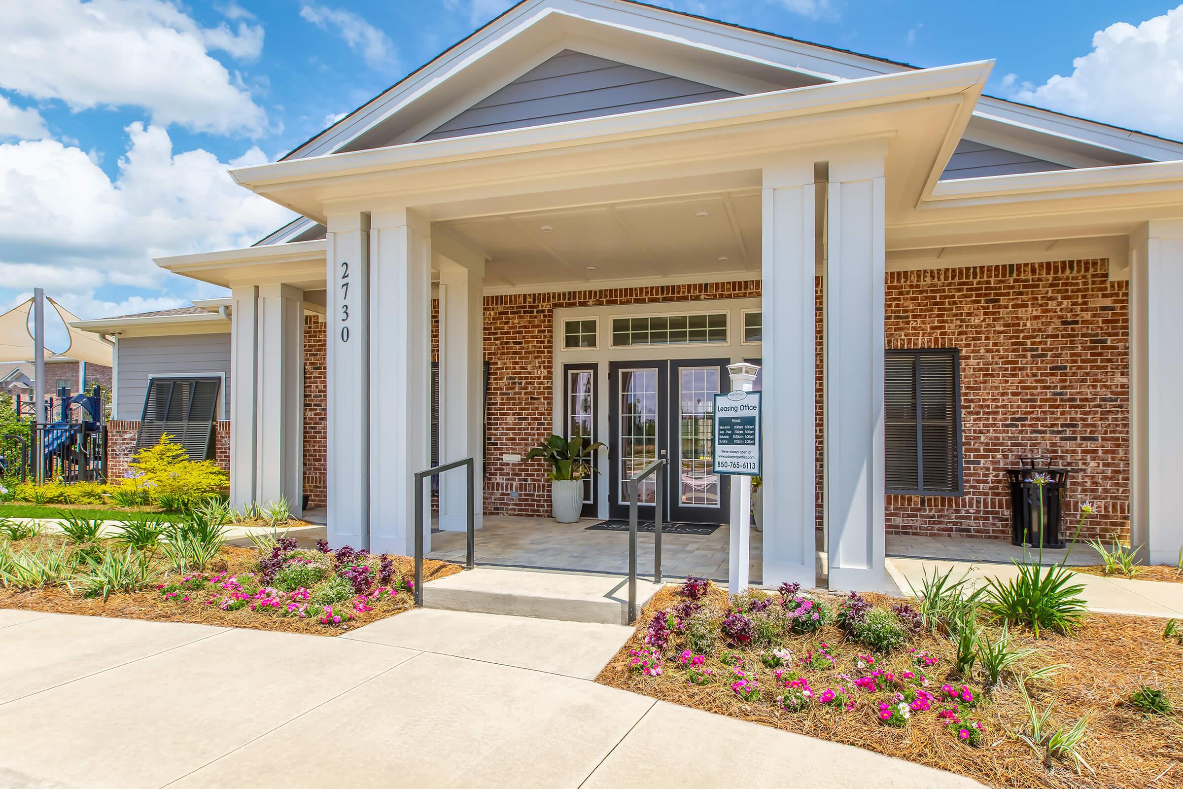 A well-lit entrance of a modern building featuring large glass doors, surrounded by landscaped gardens with colorful flowers and greenery. The facade has a combination of brick and light-colored siding, and the address "2730" is displayed prominently. Blue skies and fluffy clouds enhance the inviting atmosphere.